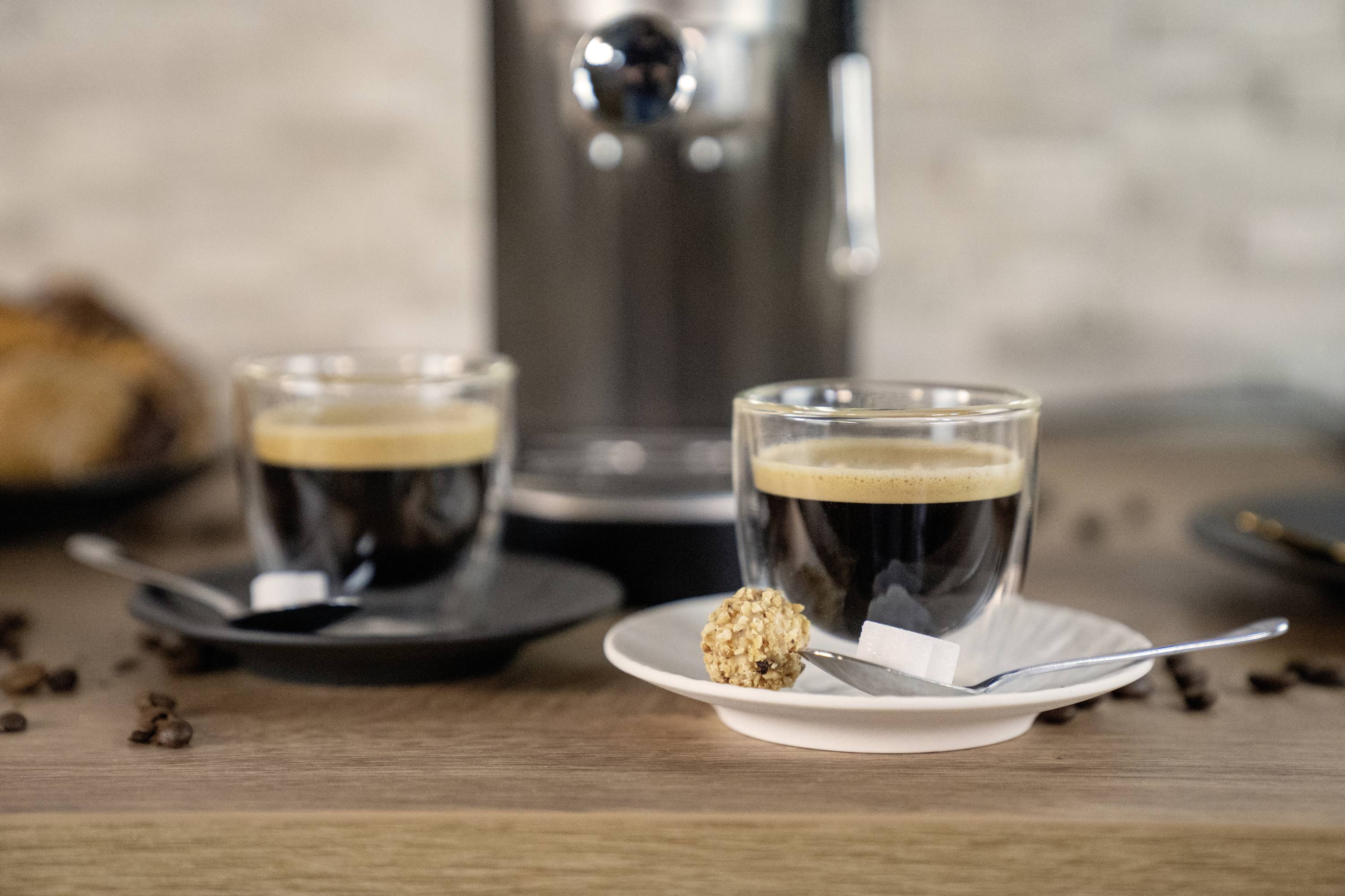 Dwie filiżanki espresso na spodeczkach z łyżeczkami, słodycz przypominająca pralinę na jednej z łyżeczek. W tle: ekspres do kawy.