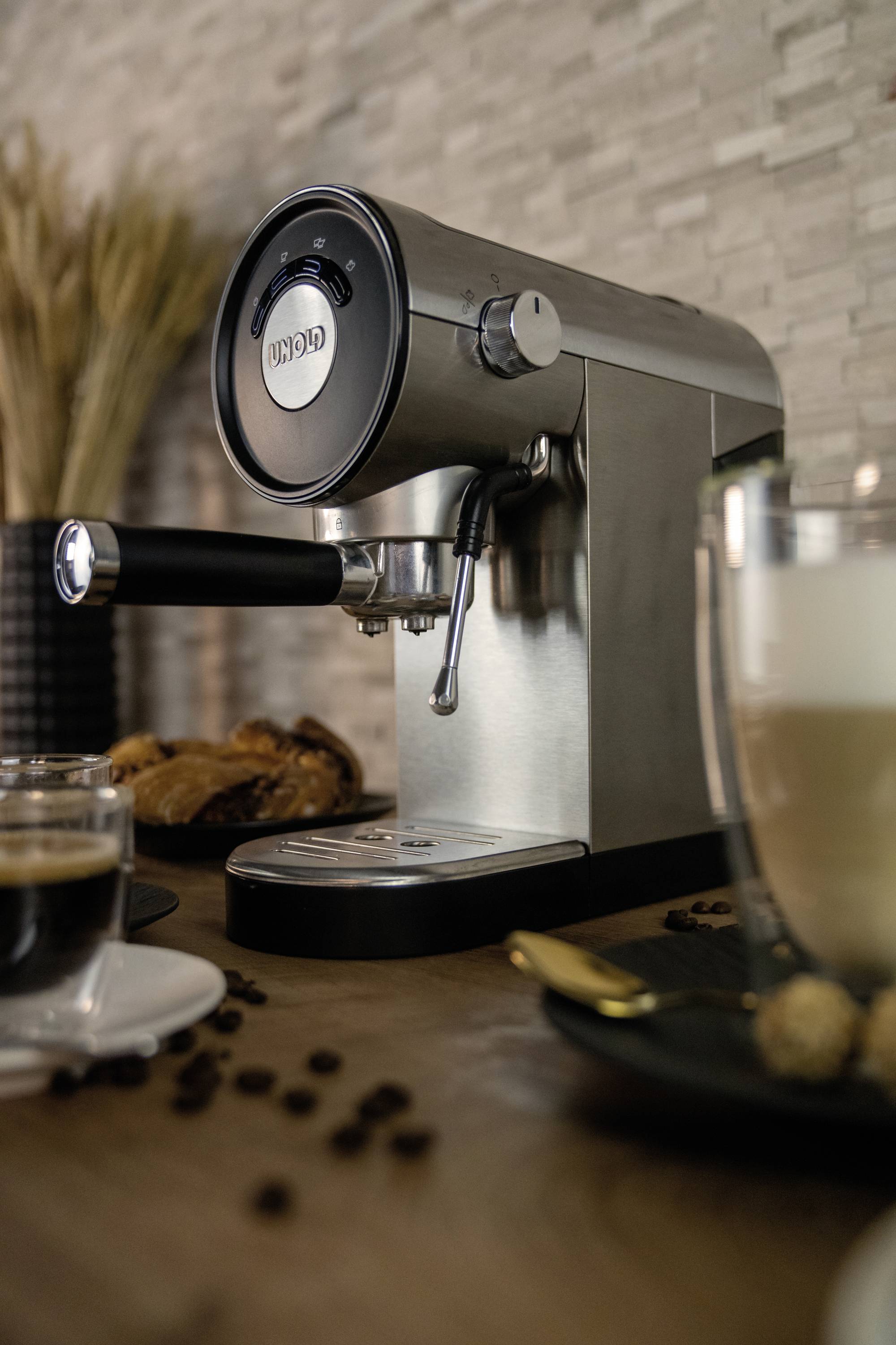 Srebrny ekspres do espresso na drewnianym blacie, otoczony filiżankami kawy, ziarnami kawy i ciastkami; tworzy przytulną atmosferę kawiarni.