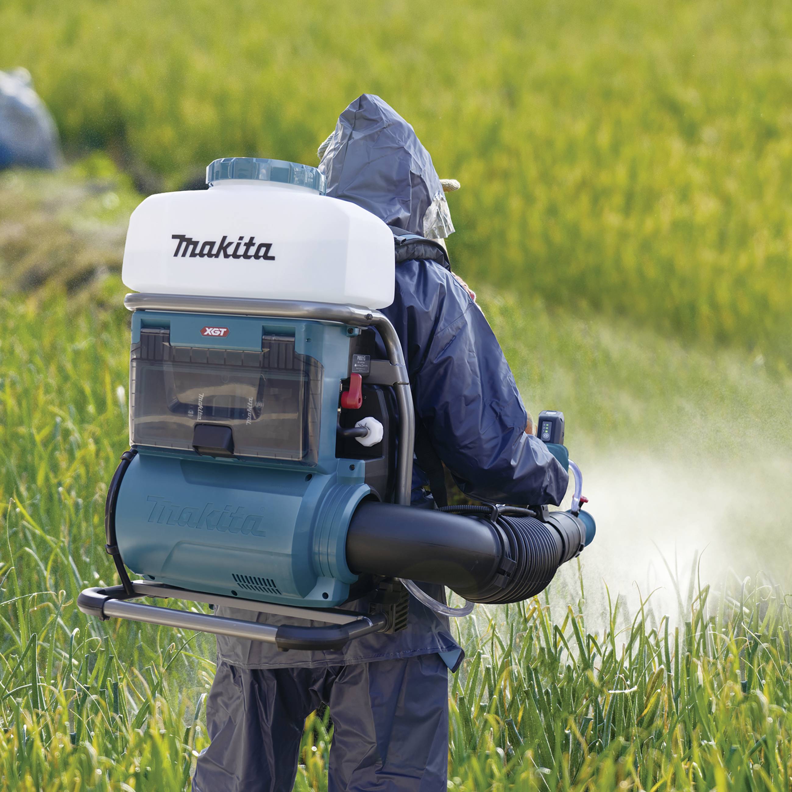 Osoba w kombinezonie ochronnym obsługuje przenośną dmuchawę do liści z białym zbiornikiem, otoczona zielonymi roślinami.