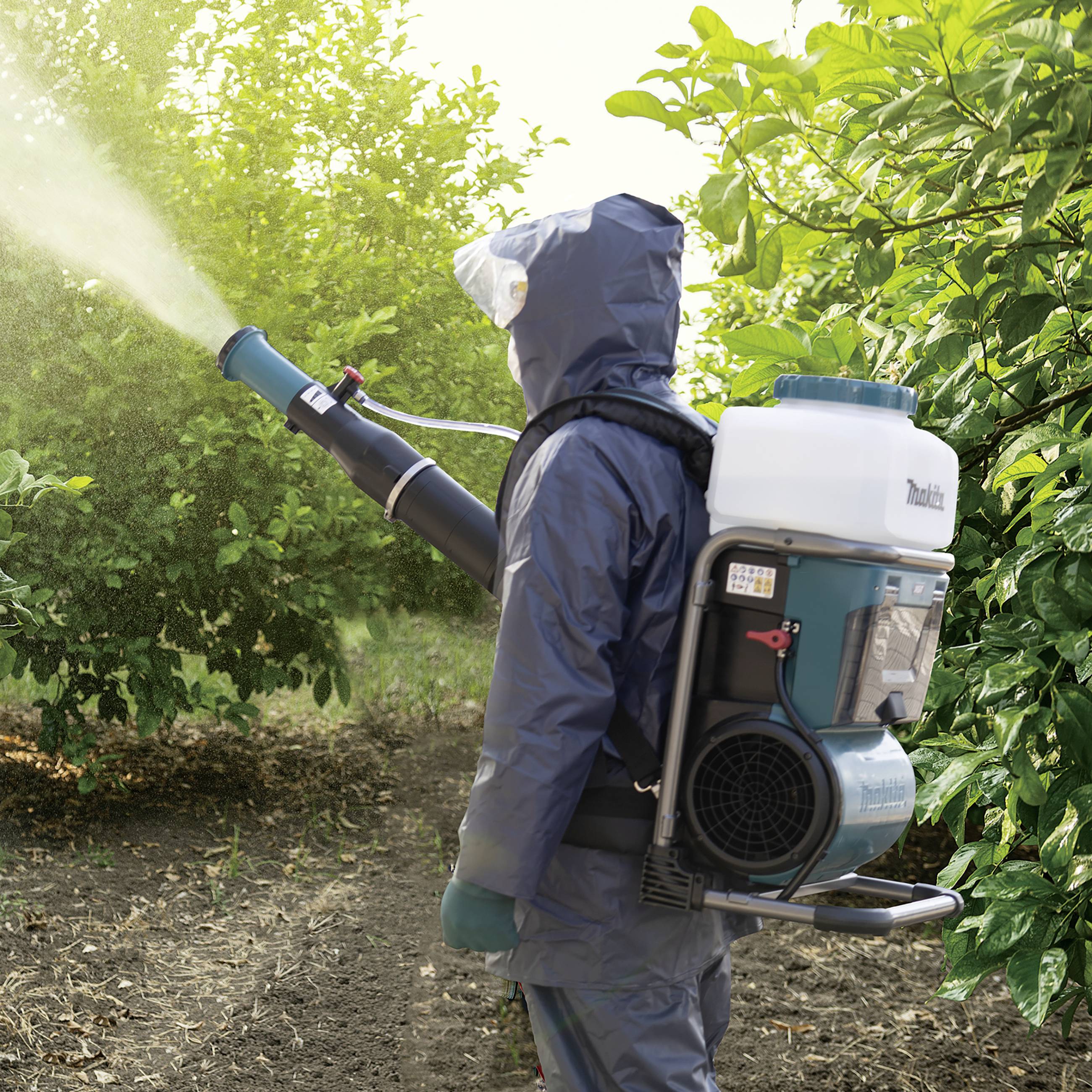 Osoba w odzieży ochronnej opryskuje rośliny w sadzie za pomocą przenośnego urządzenia.