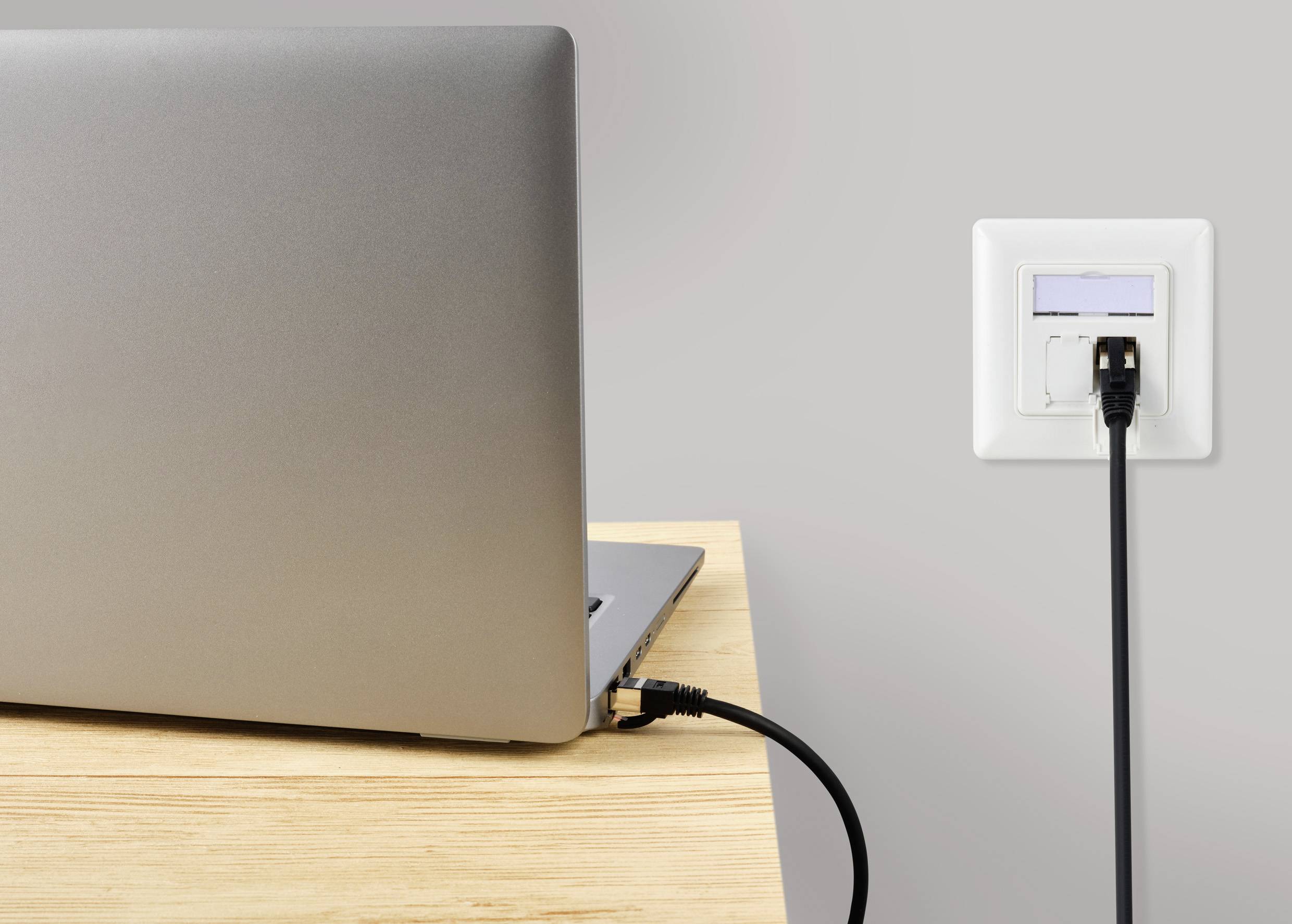 Laptop na drewnianym stole jest ładowany przez czarną ładowarkę podłączoną do białego gniazdka elektrycznego na ścianie.