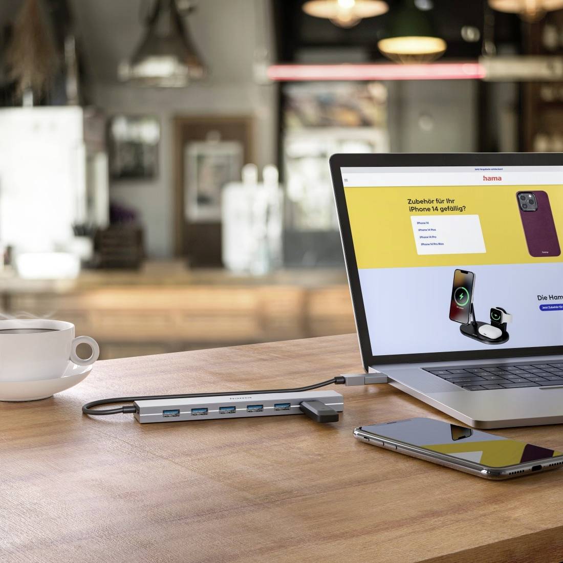 Laptop wyświetla reklamę 'Akcesoriów do iPhone 14', w tym etui i ładowarki. Obok leży smartfon, a na stole stoi filiżanka kawy.