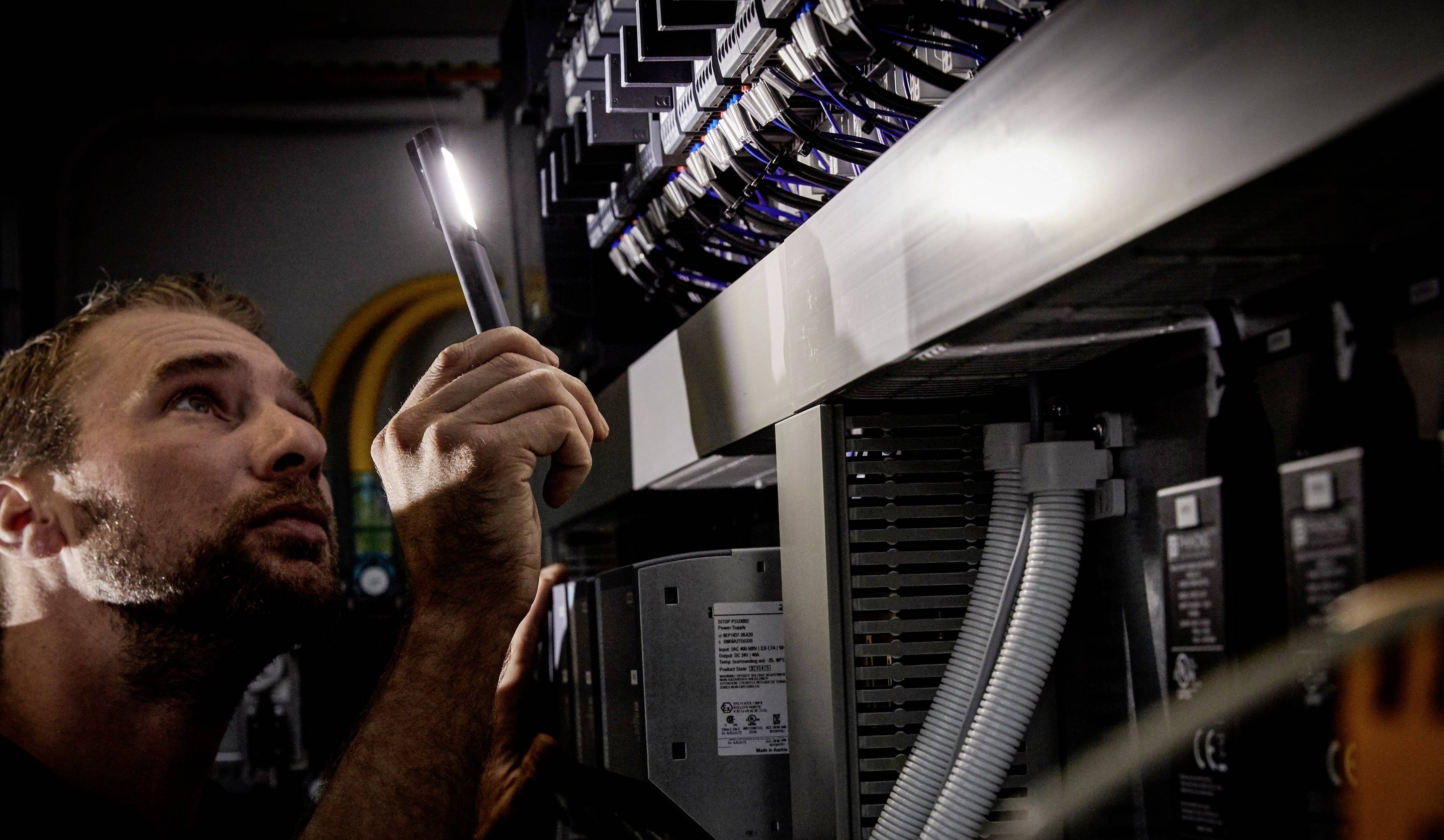 Mężczyzna sprawdza elektryczne przewody i złącza w szafce elektrycznej za pomocą latarki.