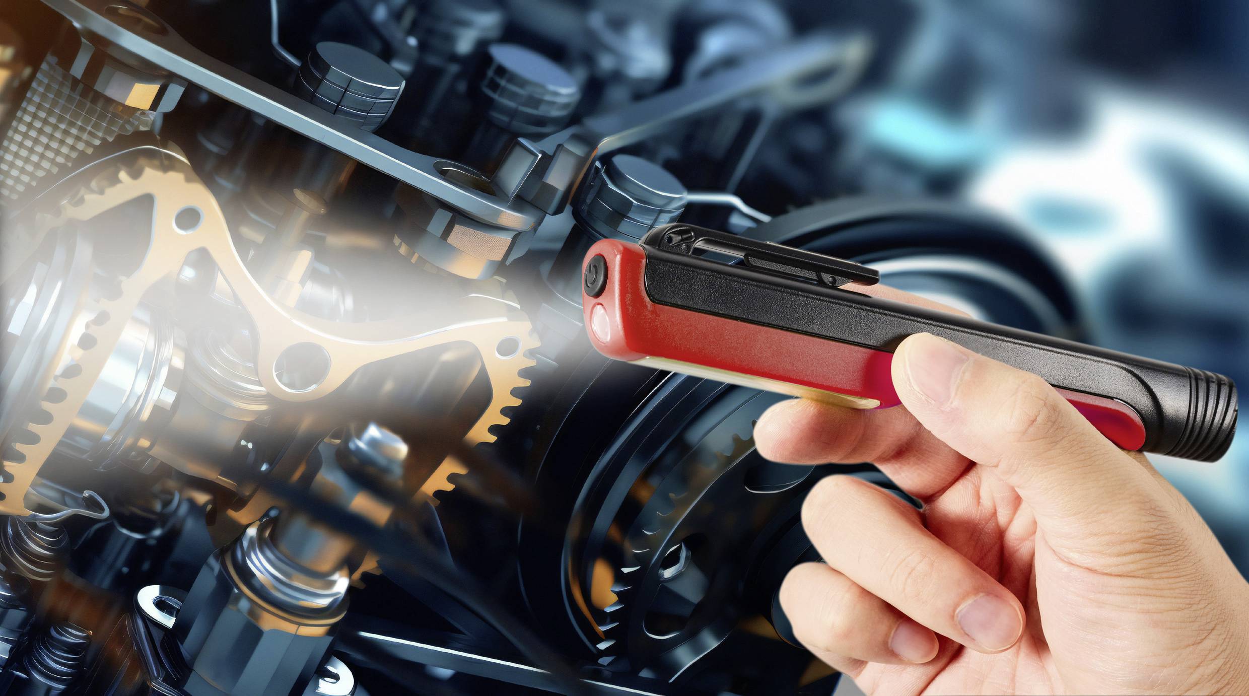 Osoba trzyma czerwono-czarne urządzenie skierowane na elementy złożonego silnika lub systemu mechanicznego.