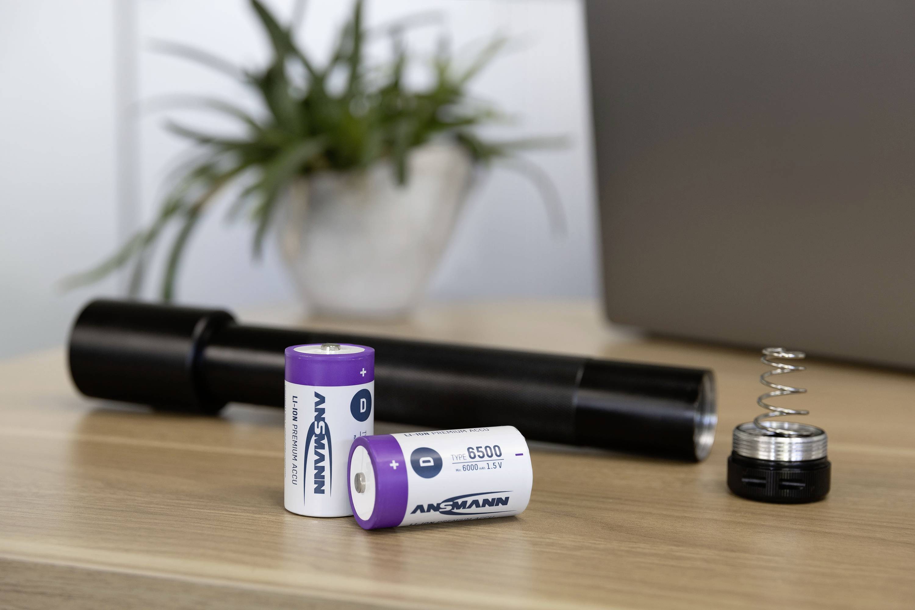 Dwie fioletowe baterie z napisem 'ANSMANN 6500' obok rozłożonej latarki na drewnianym stole.