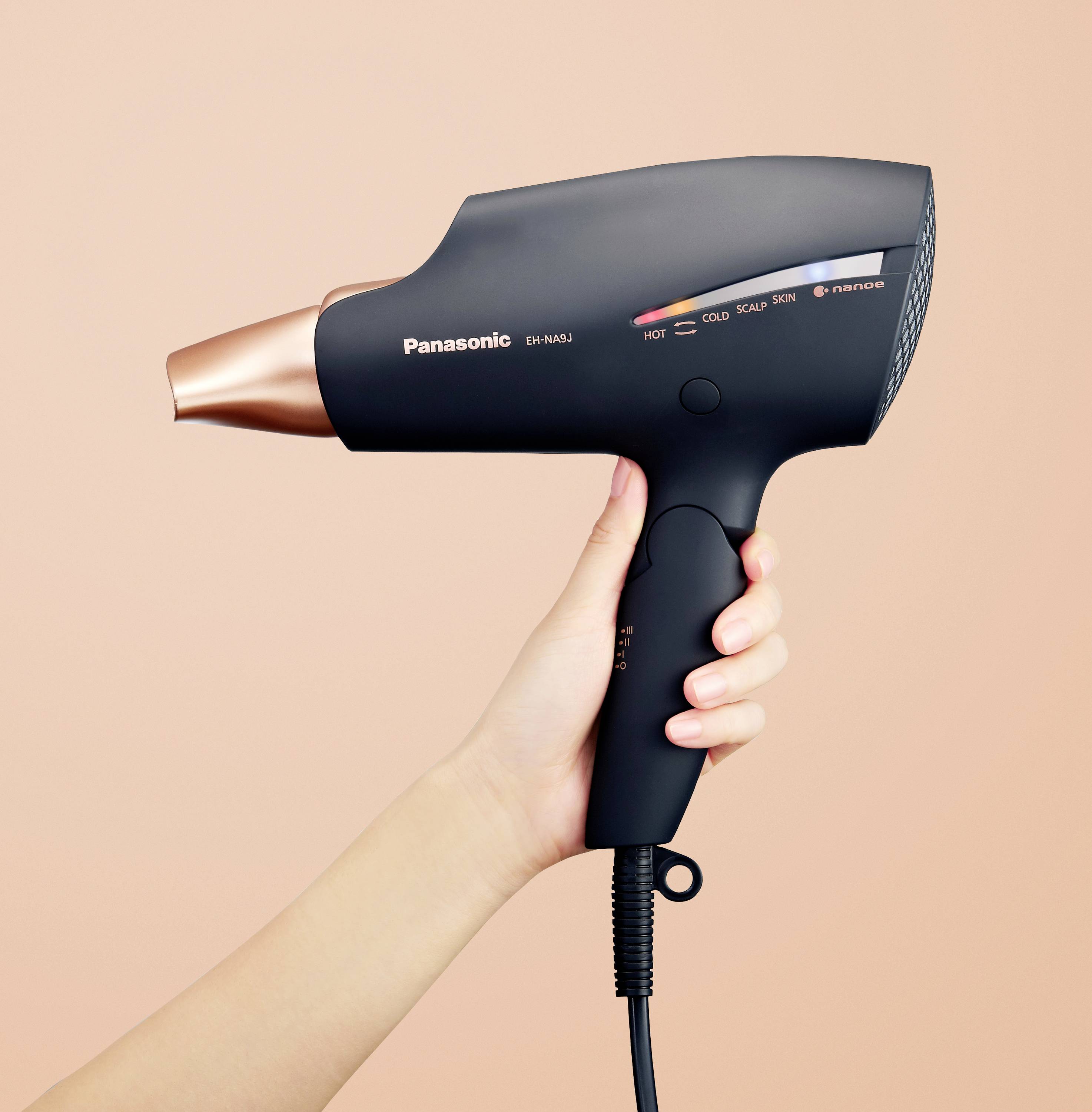 Ręka trzyma czarną suszarkę do włosów na tle w kolorze brzoskwini. Suszarka posiada ustawienia "Gorąco", "Zimno" i "Skóra".