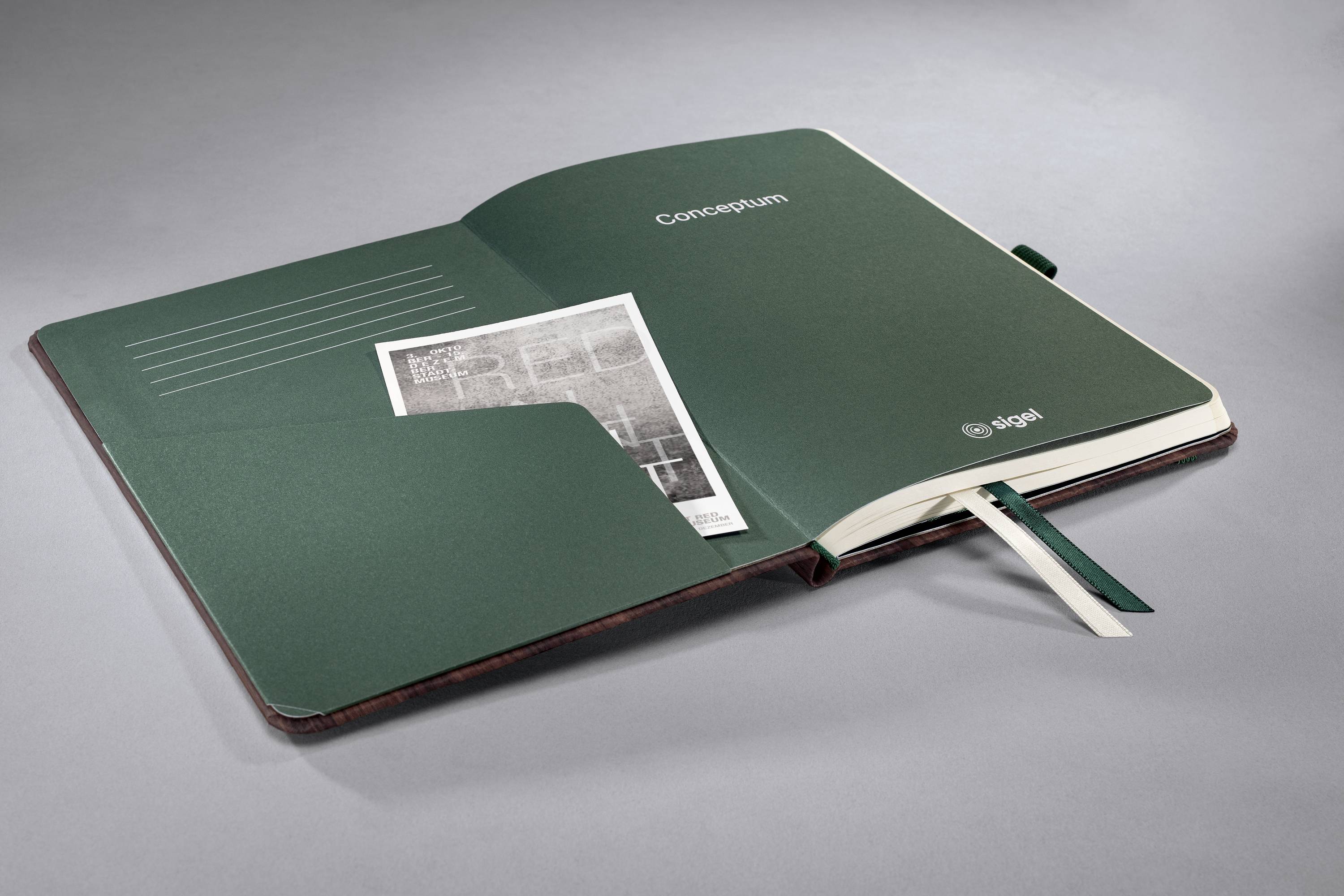 Otwarta zeszyt w zielonych okładkach. Wewnątrz w jednej z kieszeni oprawy tkwi czarno-biała strona gazety.