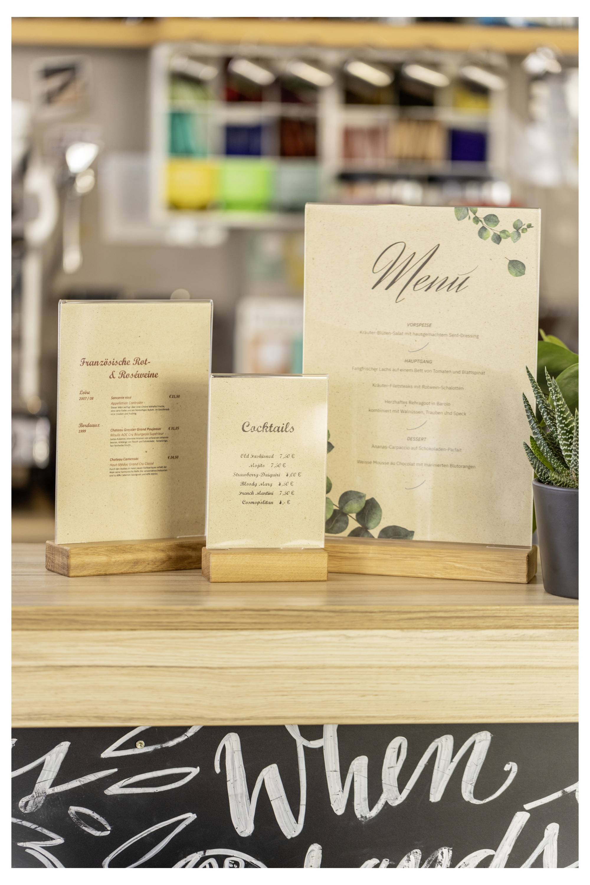 Trzy karty menu w kolorze kremowym na drewnianych podstawkach wystawione na ladorze, w tym duża karta „Menu", karta „Koktajle" i mniejsze menu.