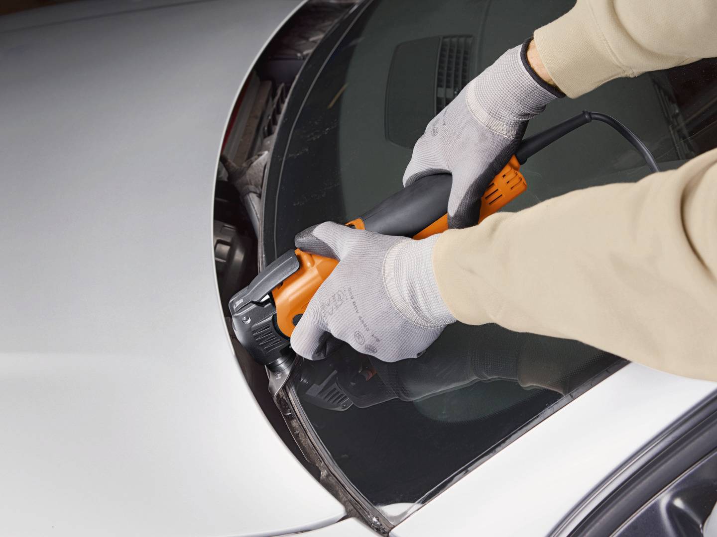 Osoba wycina przednią szybę samochodu za pomocą narzędzia elektrycznego. Ręce są zabezpieczone rękawicami ochronnymi.
