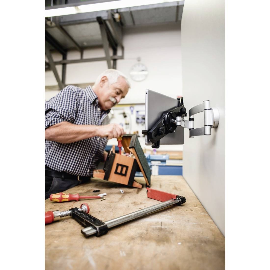 Starszy mężczyzna pracuje przy wkrętaku w warsztacie, podczas gdy tablet jest zamocowany na ścienie. Na stole leżą różne narzędzia.