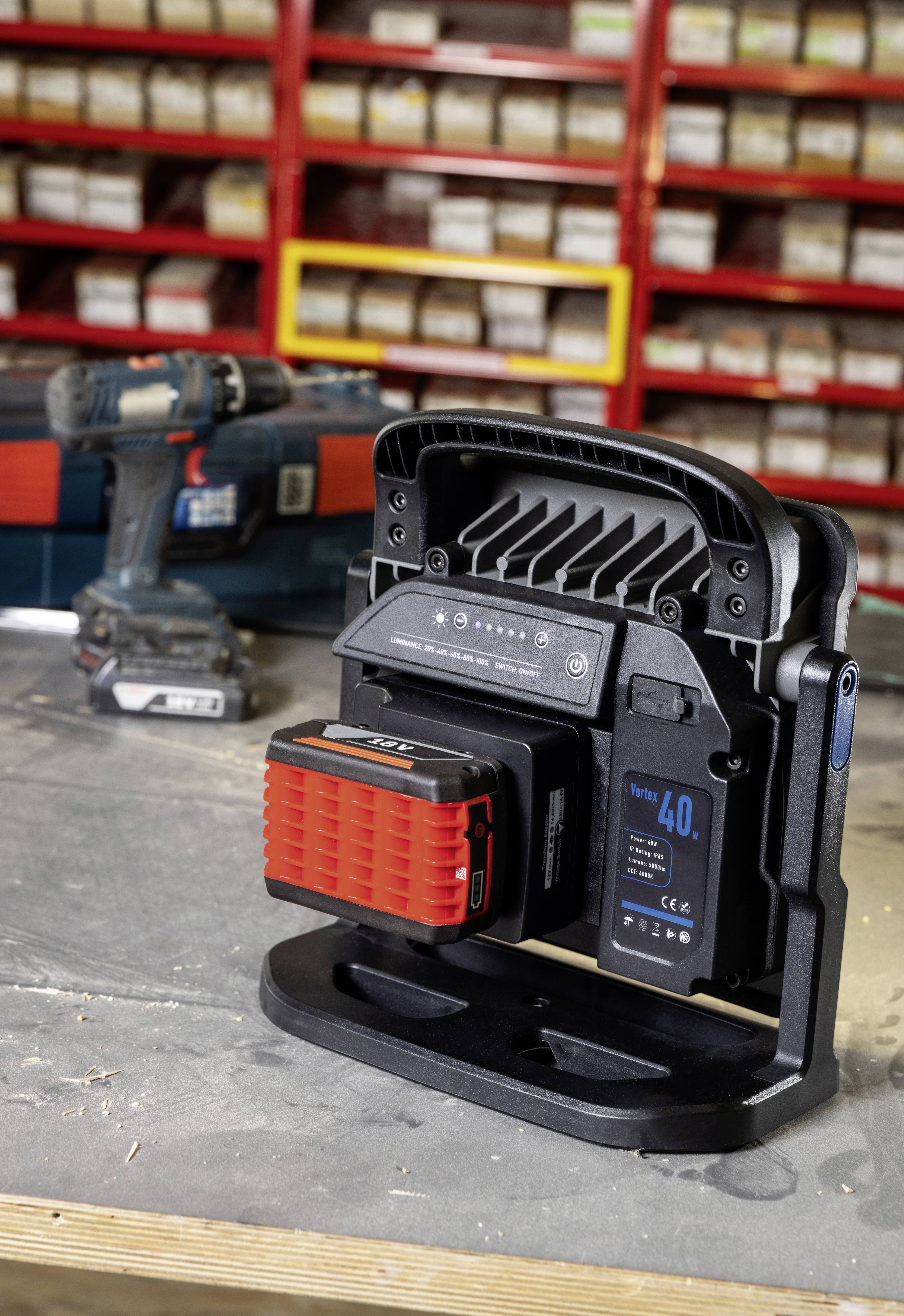 Przenośna lampa robocza LED z zamontowanym akumulatorem stoi na stole warsztatowym. W tle widoczne są regały z pudełkami oraz wiertarka.