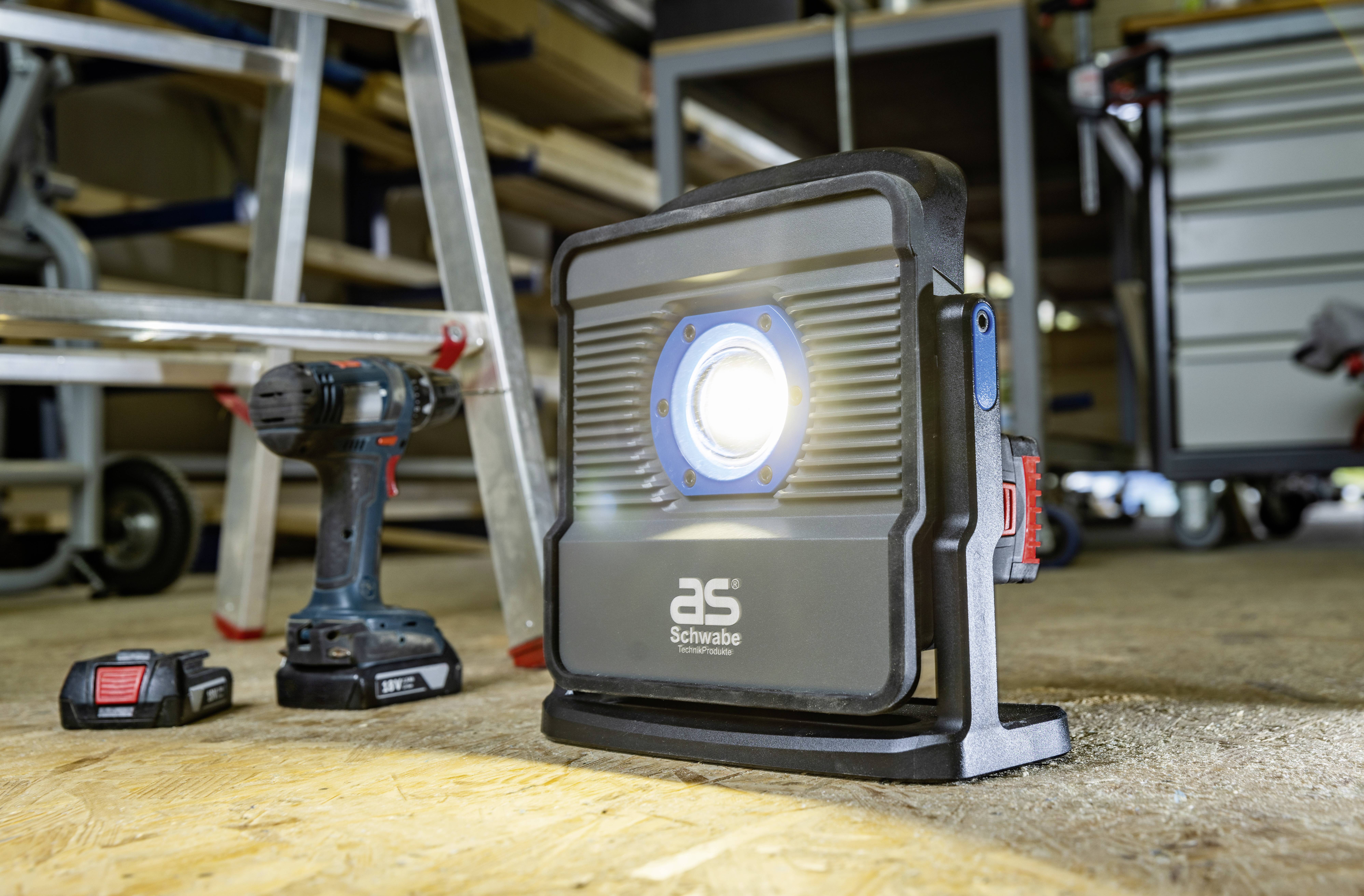 Plac budowy z lampą roboczą, wiertarką i akumulatorami na podłodze; w tle regały i narzędzia.