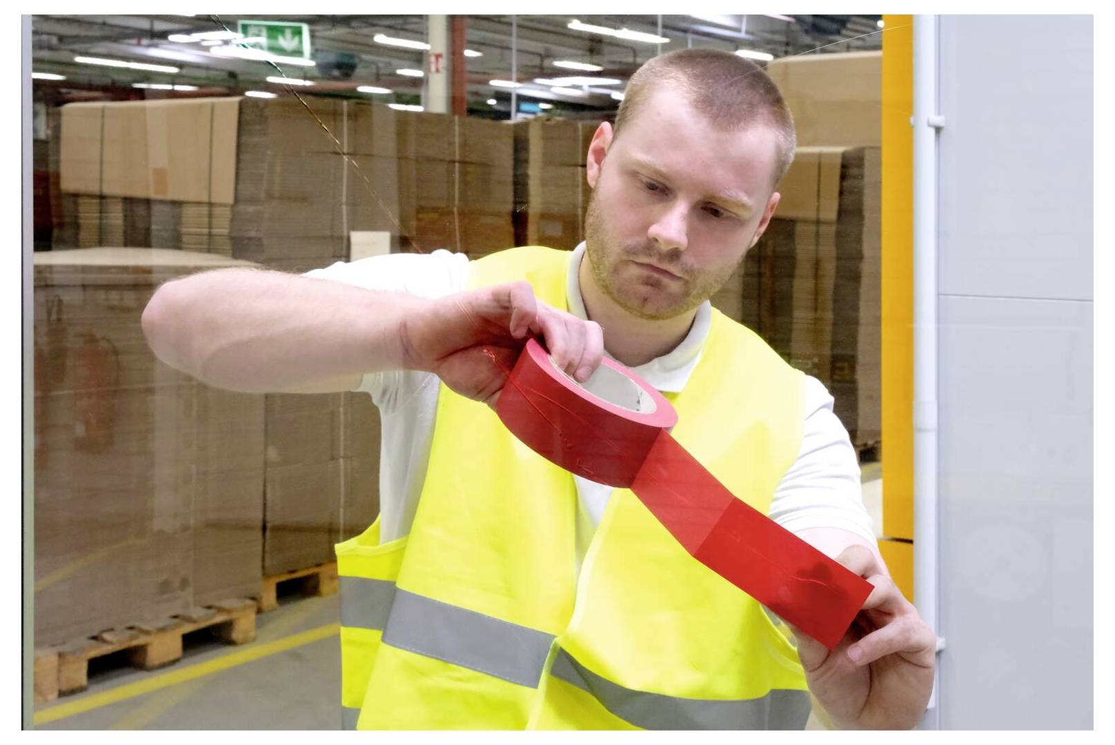 Mężczyzna w kamizelce ostrzegawczej nakłada czerwoną taśmę na szklaną powierzchnię w magazynie, w tle widać stosy kartonowych pudełek.