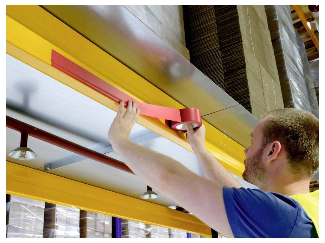 Osoba w magazynie nakłada czerwoną taśmę na metalową belkę pod regałami, z widocznymi stosami kartonu powyżej.