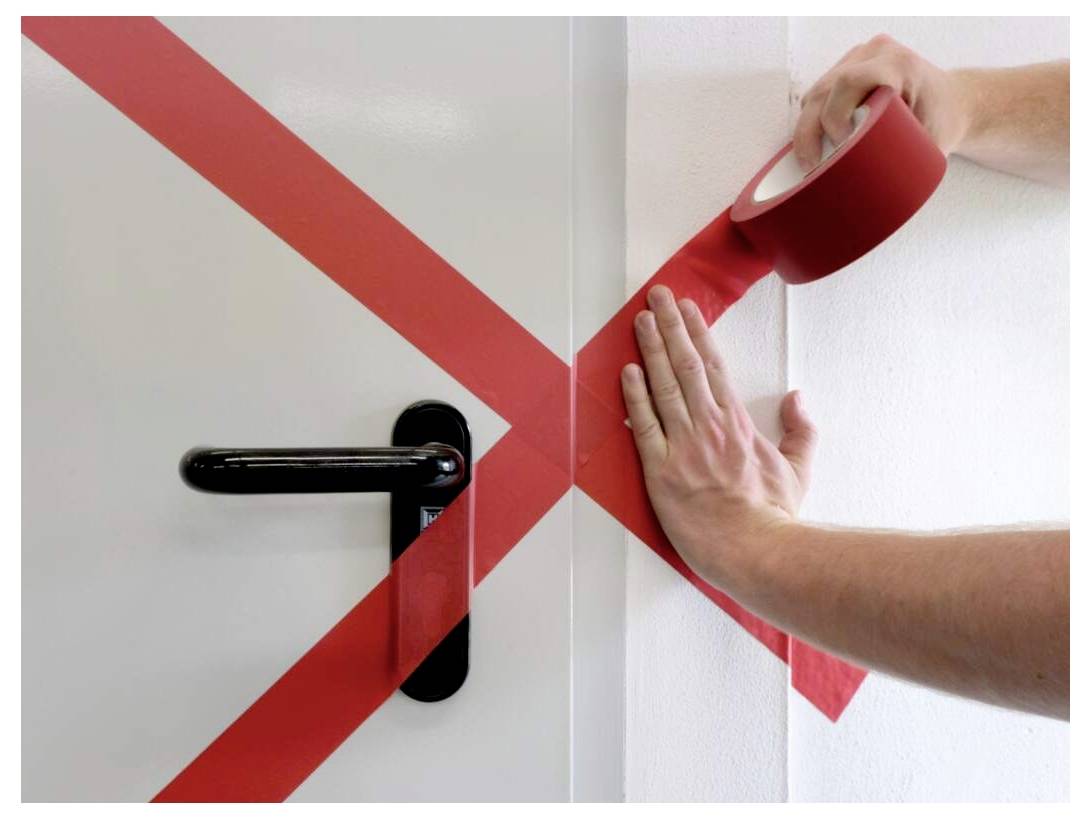 Osoba nakładająca czerwoną taśmę w kształcie litery 'X' na drzwi i ścianę, zakrywająca klamkę drzwi.