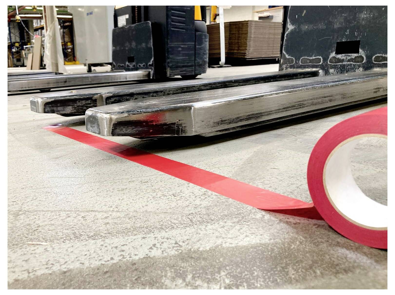 Zbliżenie wózka widłowego z widłami ustawionymi równolegle nad czerwoną taśmą podłogową w magazynie, podkreślające precyzyjne ustawienie dla zachowania bezpieczeństwa.