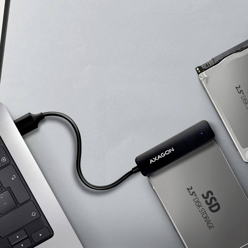 Laptop jest podłączony kablem do zewnętrznego dysku SSD o rozmiarze 2,5 cala. Dysk SSD leży obok laptopa na szarym stole.