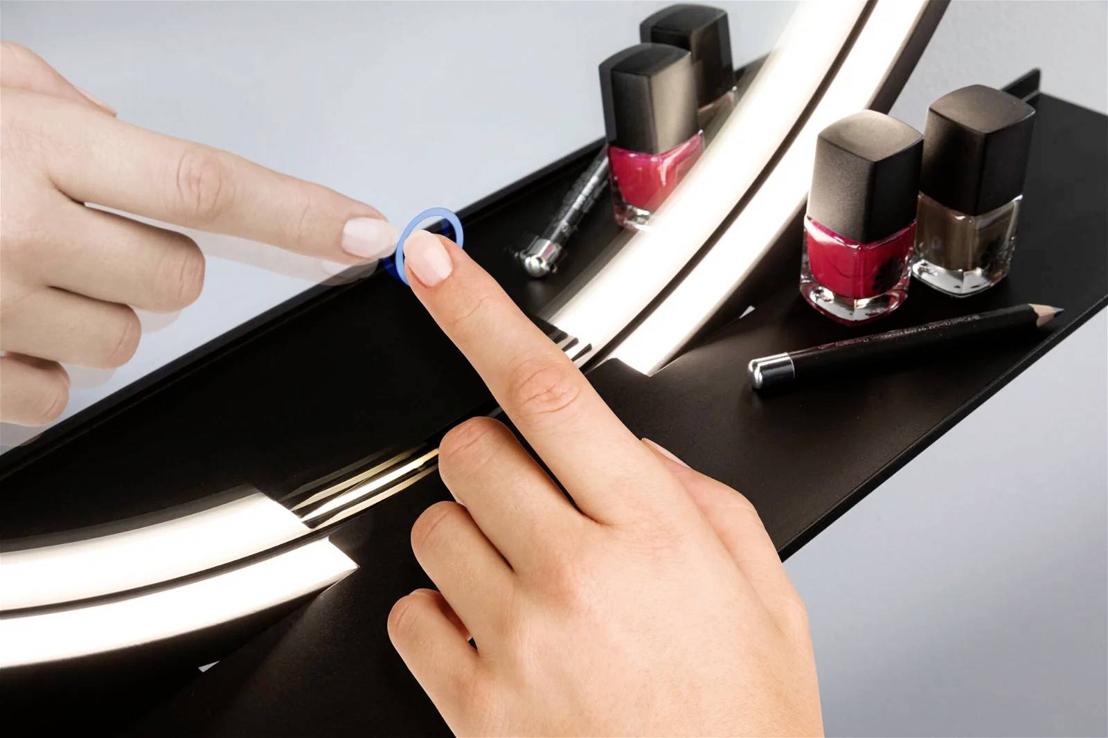 Dwie dłonie z lakierem do paznokci, odbicie w lustrze, różne kolory lakieru w tle, jedna dłoń nakłada czerwony lakier do paznokci.