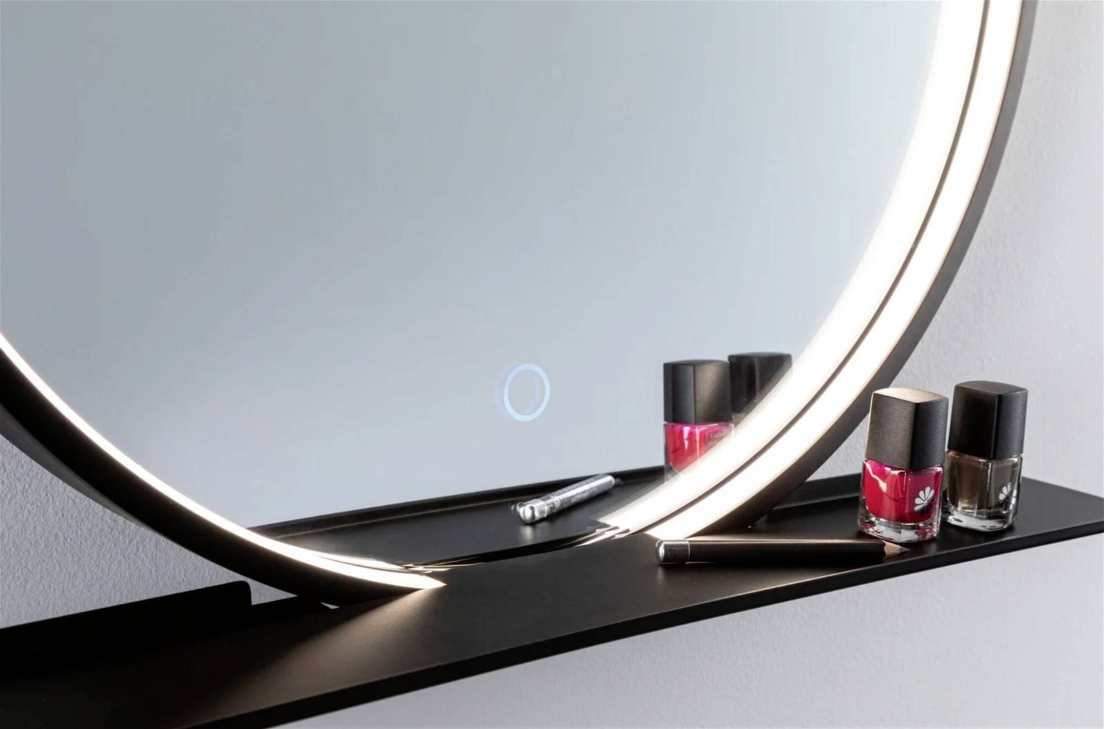 Okrągłe lustro z podświetleniem LED, ozdobione czerwonymi i czarnymi buteleczkami lakieru do paznokci na półce poniżej.