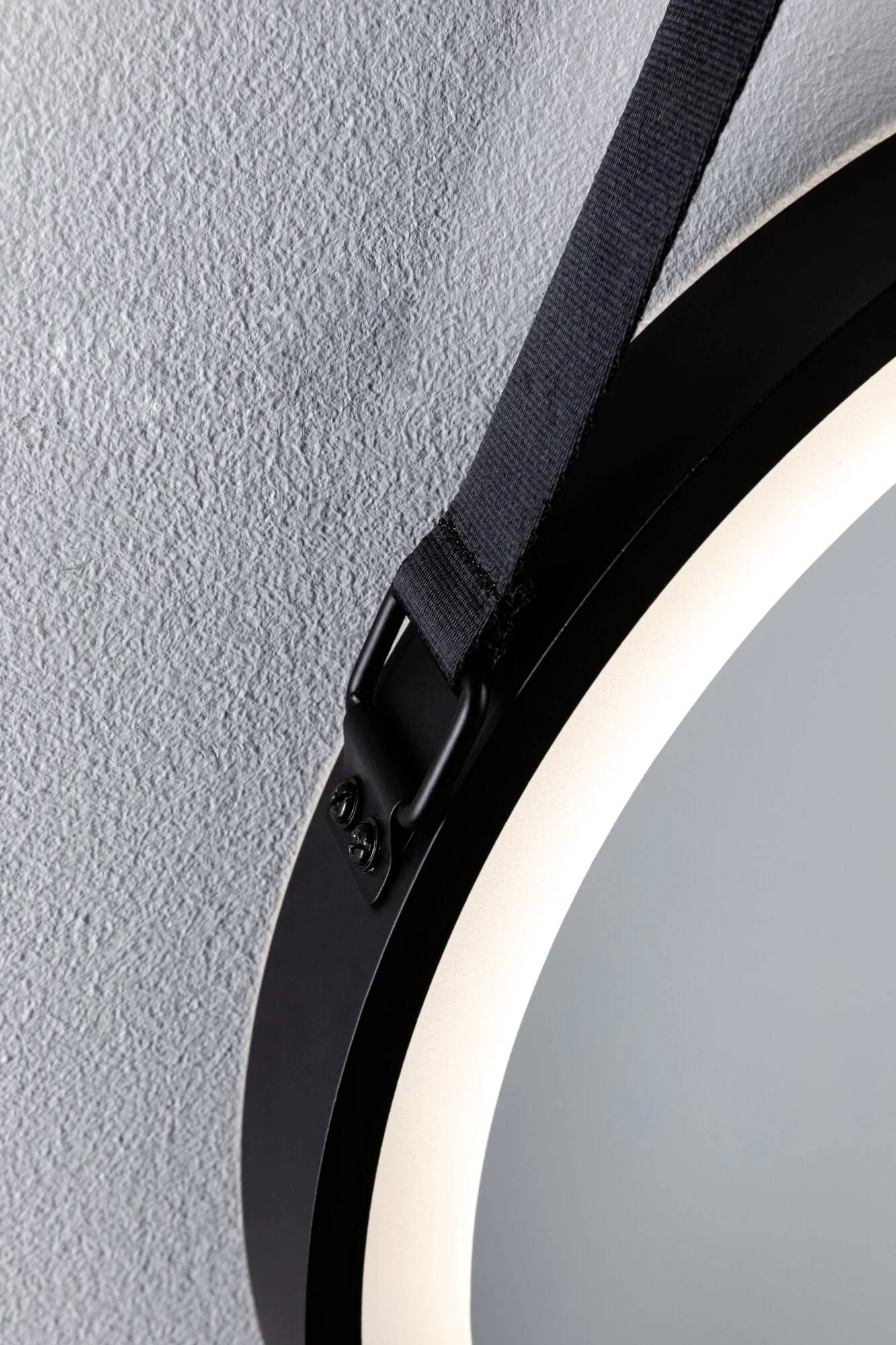 Zbliżenie na okrągłe lustro zawieszone na niebieskiej ścianie czarnym pasem. Widoczne w lewym górnym rogu obrazu.