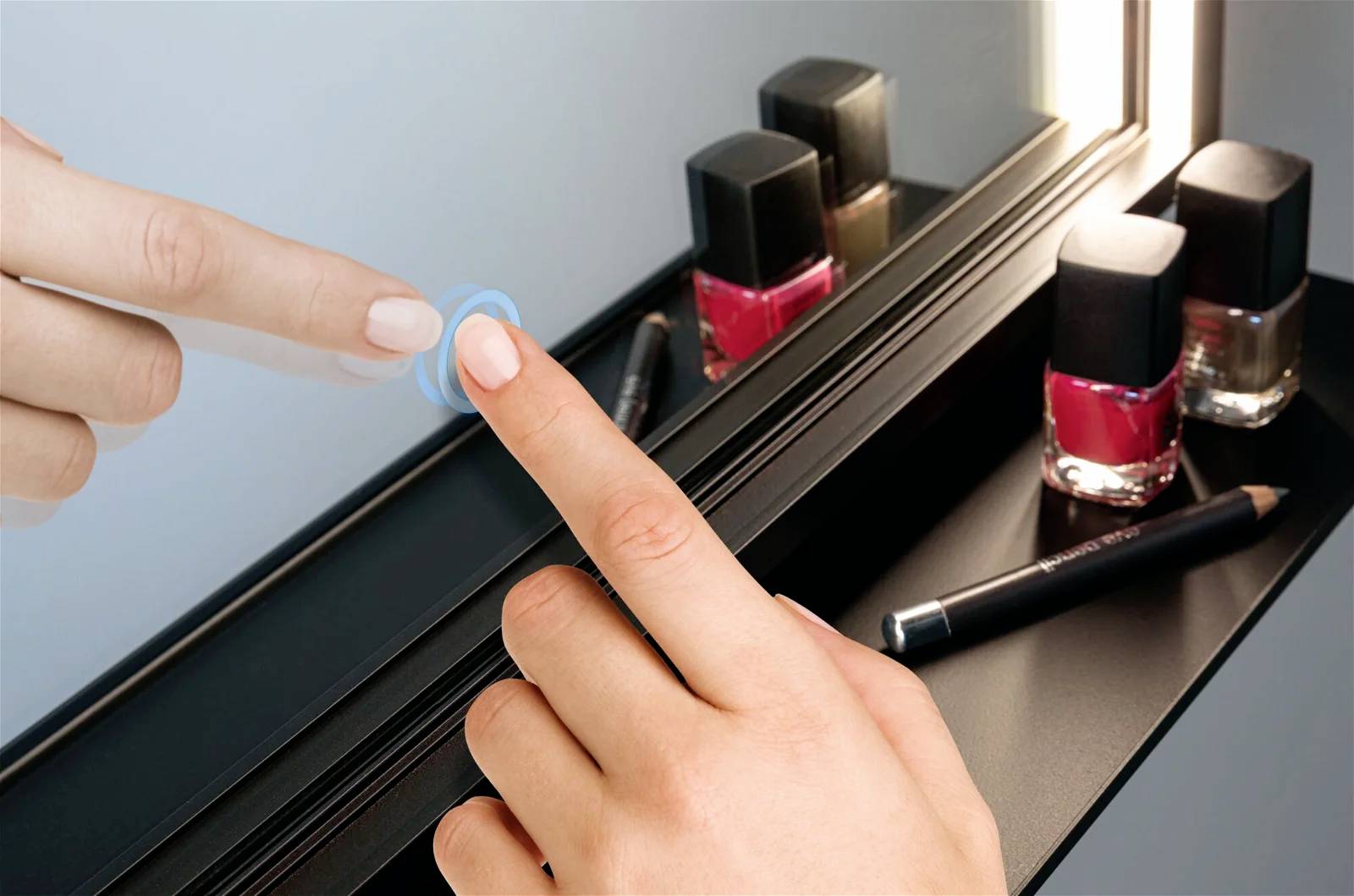 Osoba dotyka podświetlonego lustra. Na półce poniżej stoją lakiery do paznokci w odcieniach czerwieni i nude oraz czarny kredka do oczu.