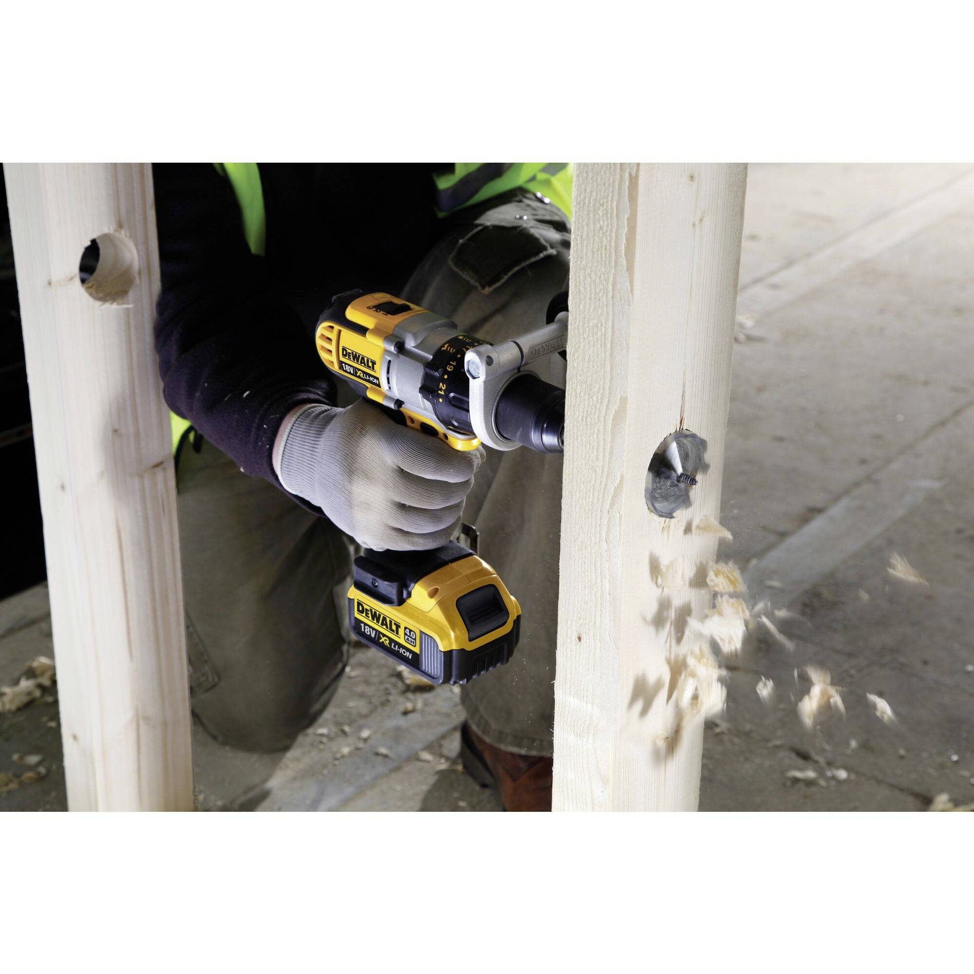 Osoba wierci akumulatorowym wiertłem otwór w belce drewnianej, a wióry drewniane lecą. W tle widoczne jest środowisko budowy.