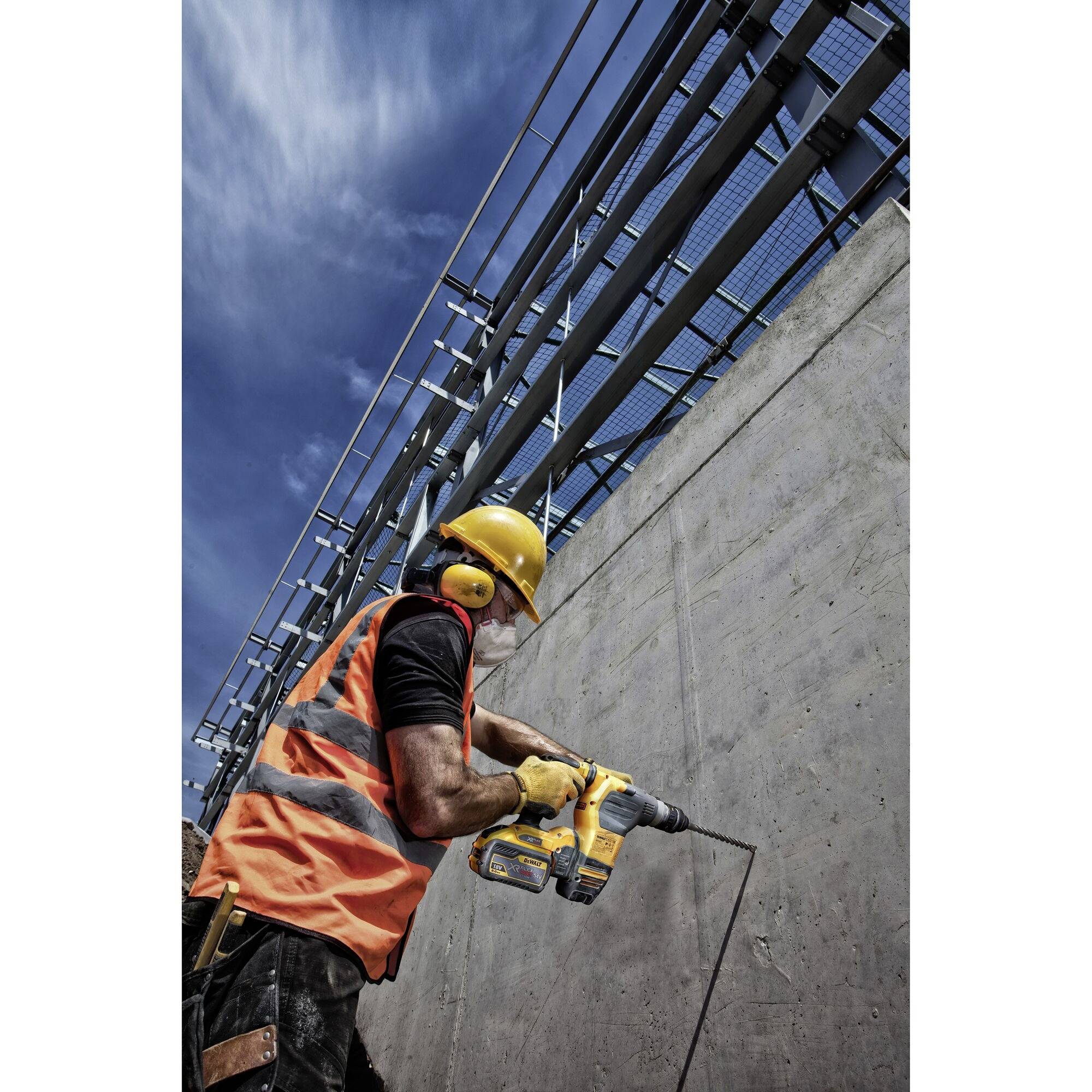 Robotnik budowlany w kasku ochronnym i kamizelce odblaskowej wierca w betonową ścianę za pomocą maszyny pod błękitnym niebem, w tle widoczny ogrodzenie zabezpieczające.