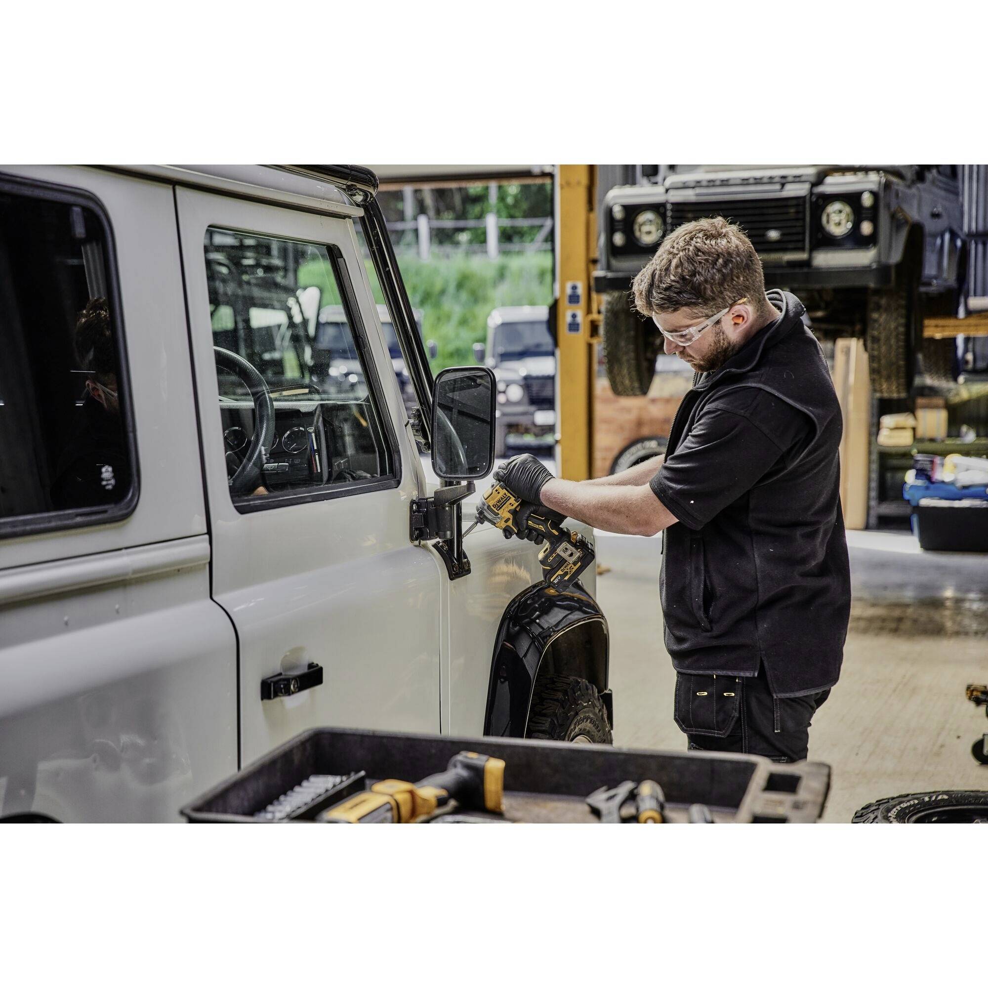 Mężczyzna skupionym wzrokiem naprawia lusterko boczne białego samochodu terenowego w warsztacie. Na pierwszym planie widoczne są narzędzia.