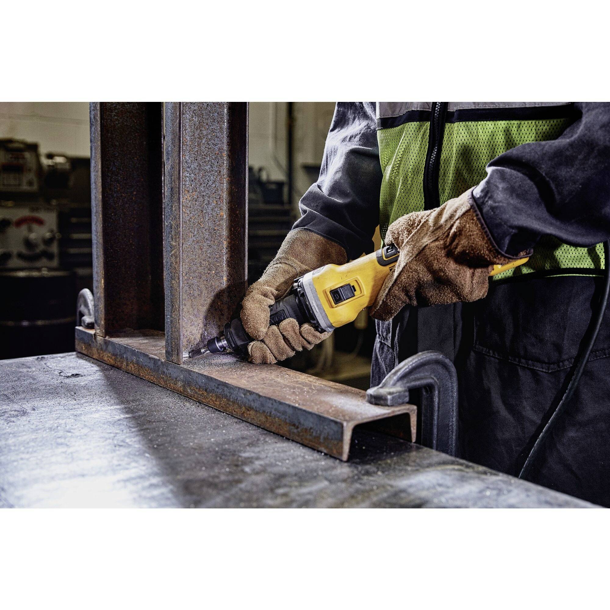 Osoba pracuje żółtą szlifierką kątową na elemencie metalowym w warsztacie. Jest ubrana w kombinezon ochronny i rękawice.