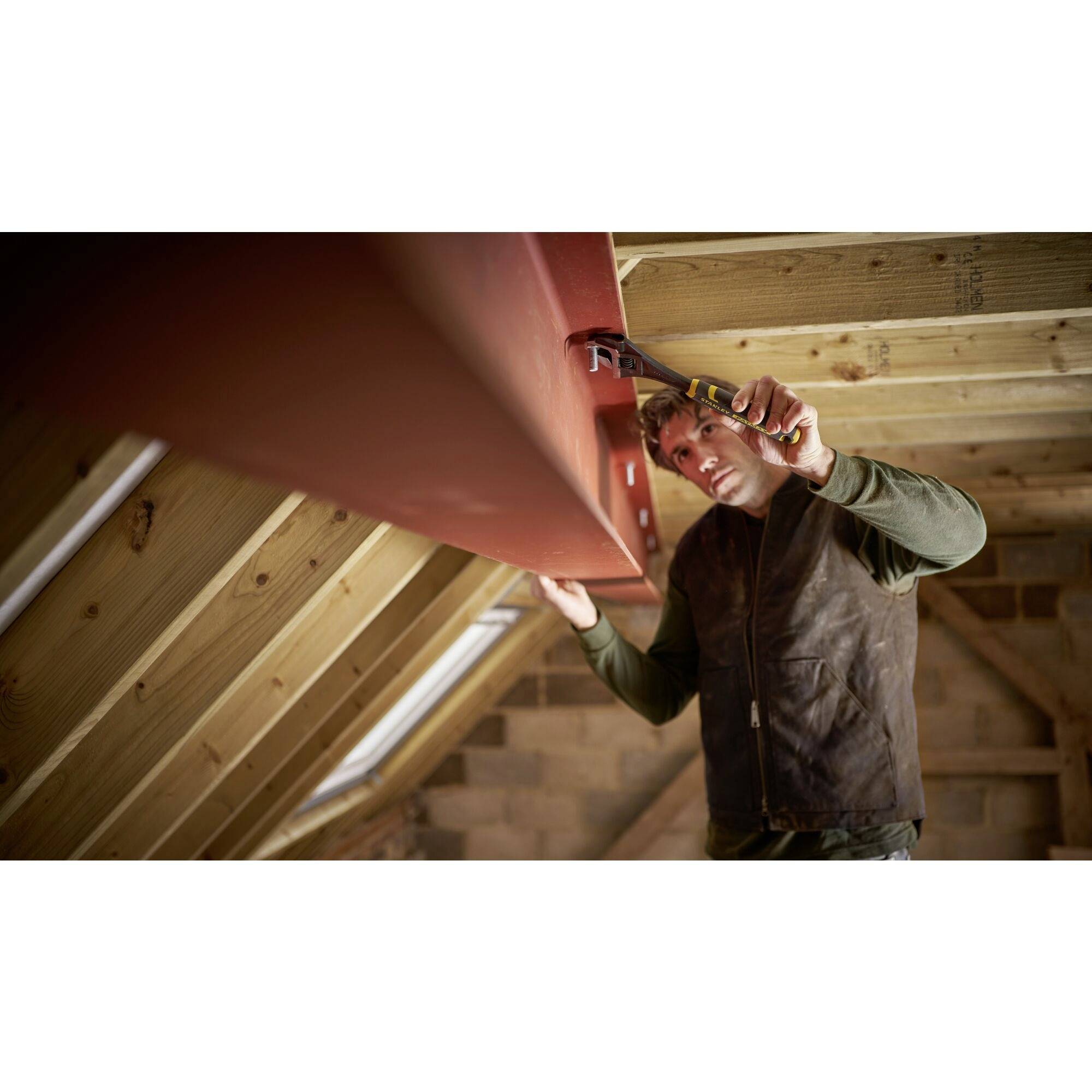 Mężczyzna w odzieży roboczej przykręca narzędziem element drewnianej konstrukcji pod więźbą dachową.