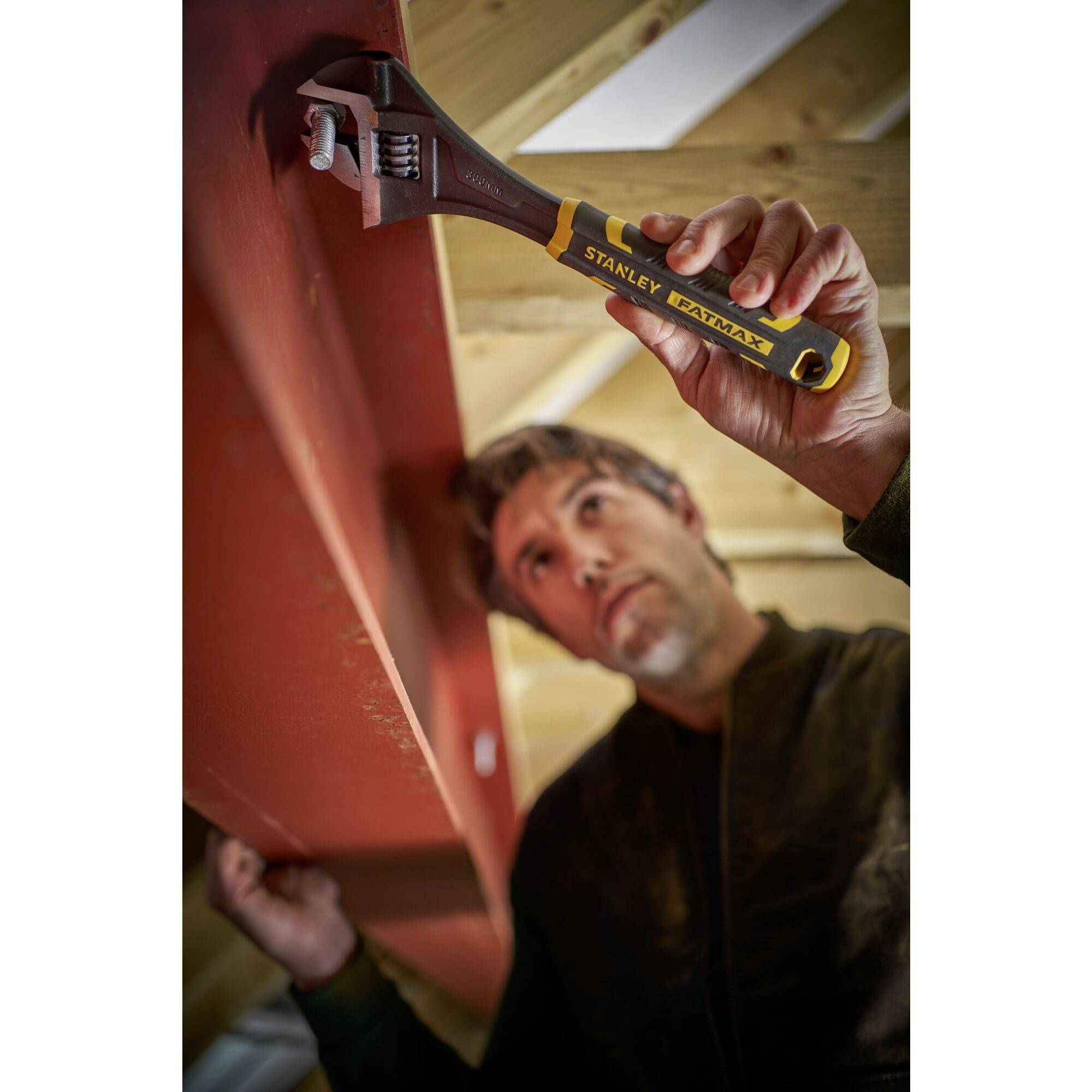 Mężczyzna pracuje za pomocą regulowanego klucza nastawnego na czerwonym stalowym elemencie nośnym w budynku z widocznymi strukturami drewnianymi.