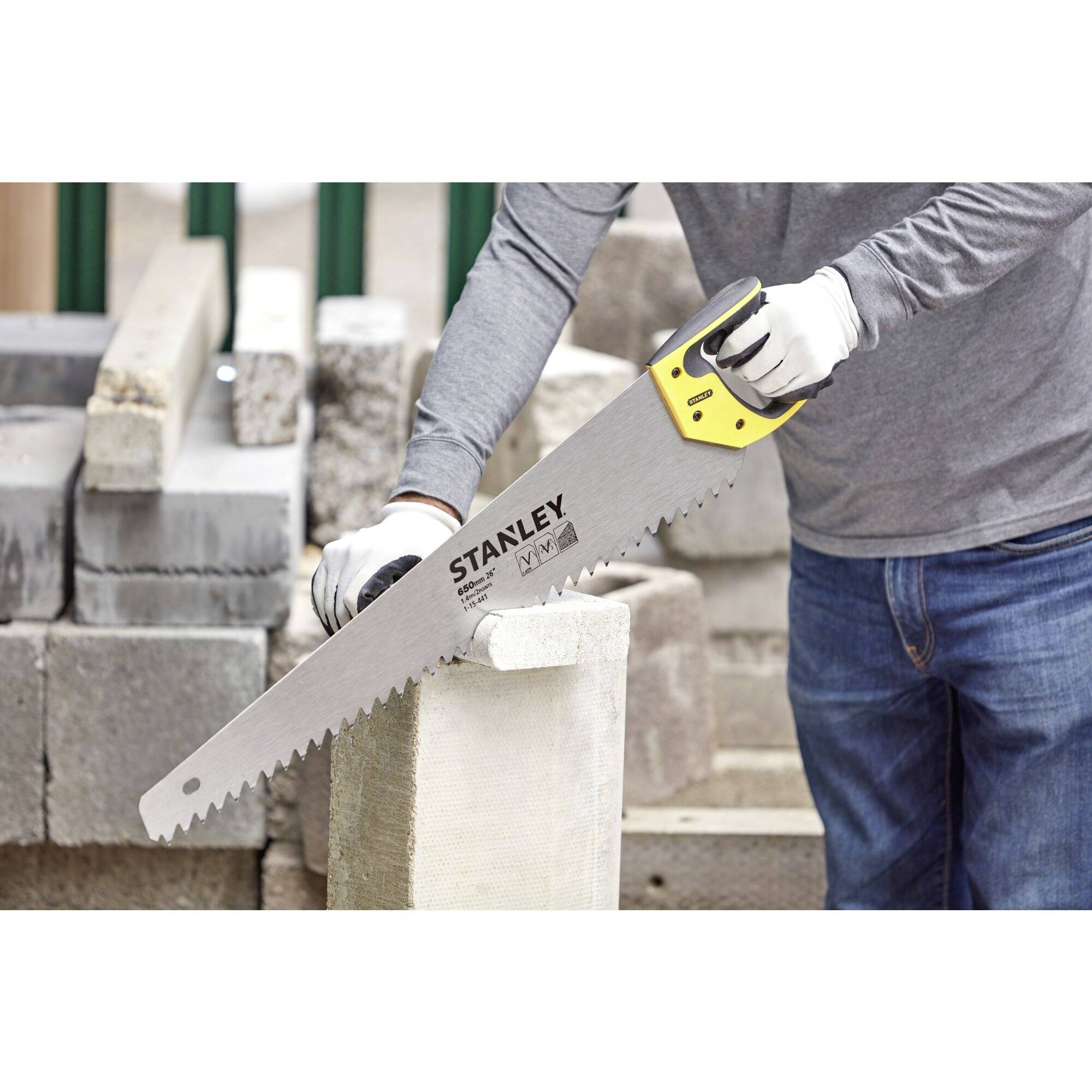 Osoba przecina blok betonowy ręczną piłą. W tle stos betonowych kostek, osoba ubrana w szarą bluzę i rękawice.