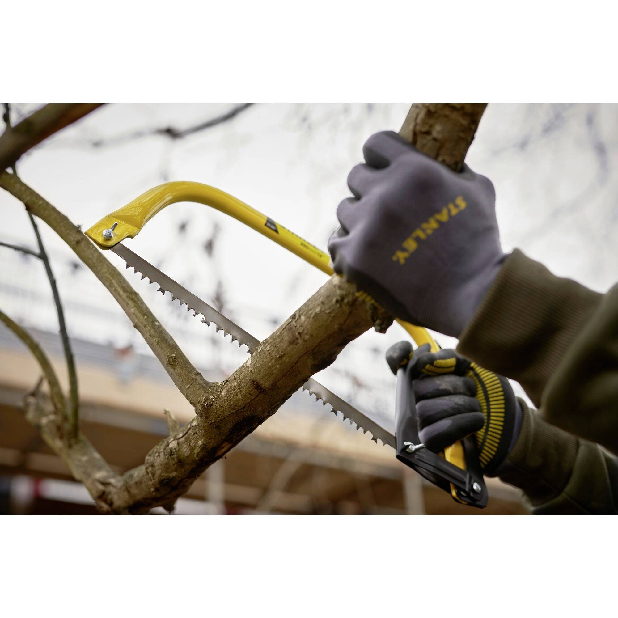 Osoba piłuje gałąź ręczną piłą. Ma na rękach rękawice i pracuje na świeżym powietrzu.