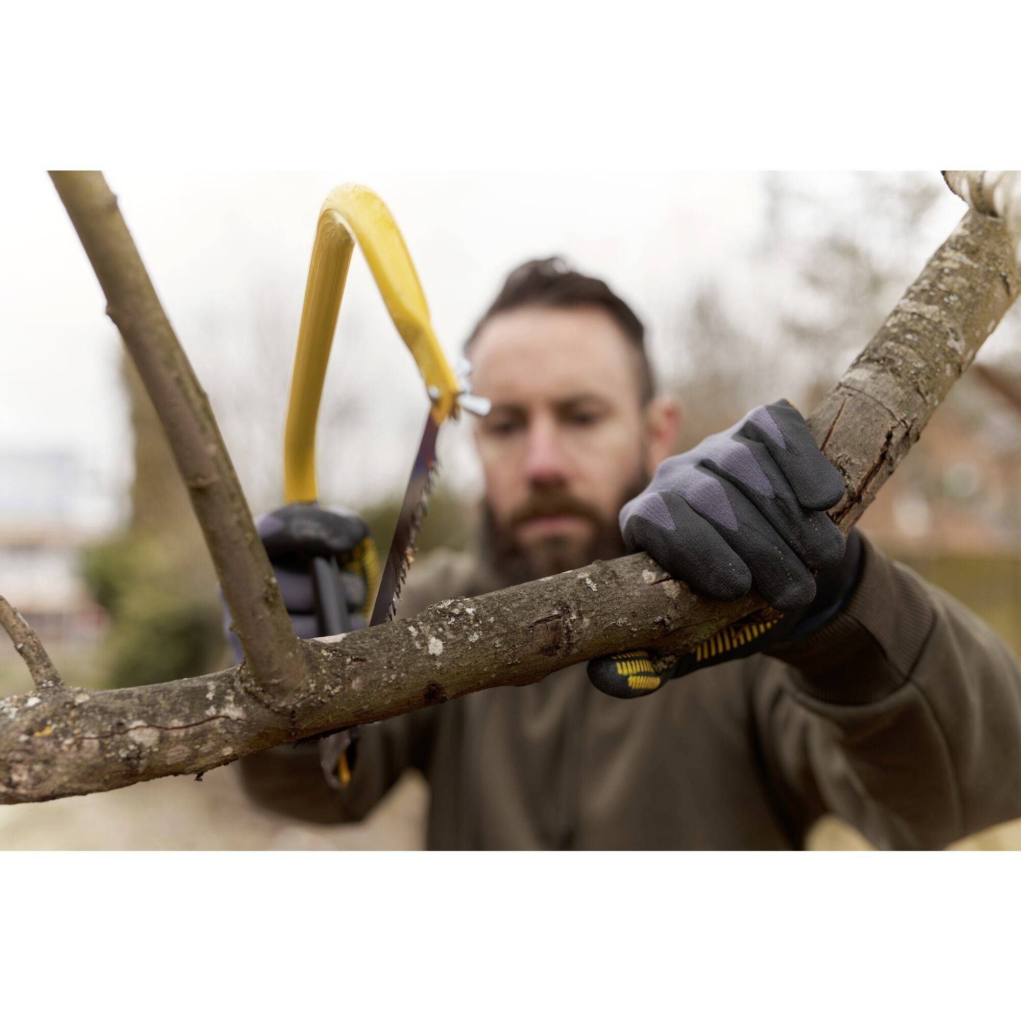 Osoba piłuje gałąź ręczną piłą na dworze. Ma na rękach rękawice. Tło jest rozmyte.