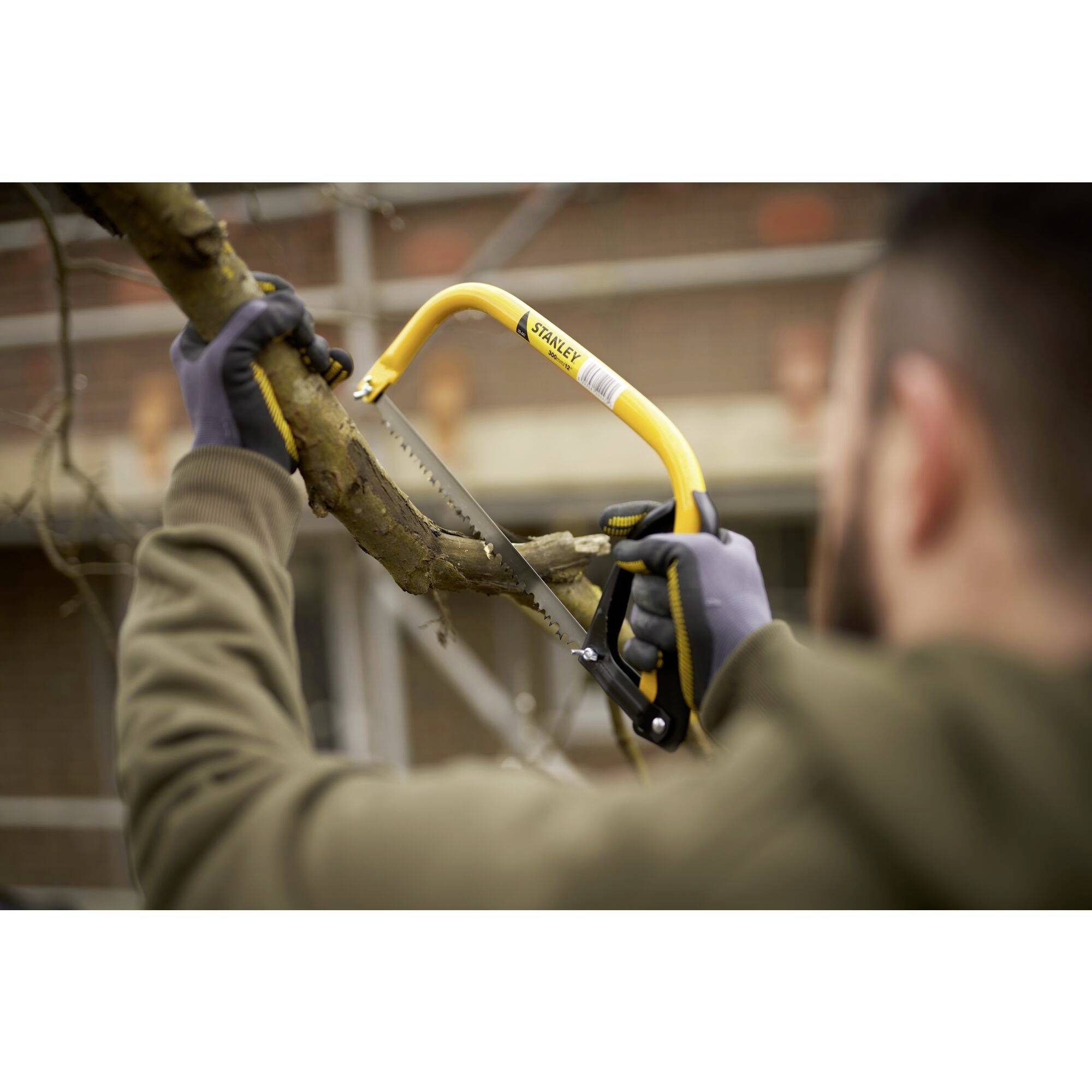 Osoba piłuje gałąź drzewa ręczną piłą. Piła ma żółty uchwyt. W tle widać budynek.