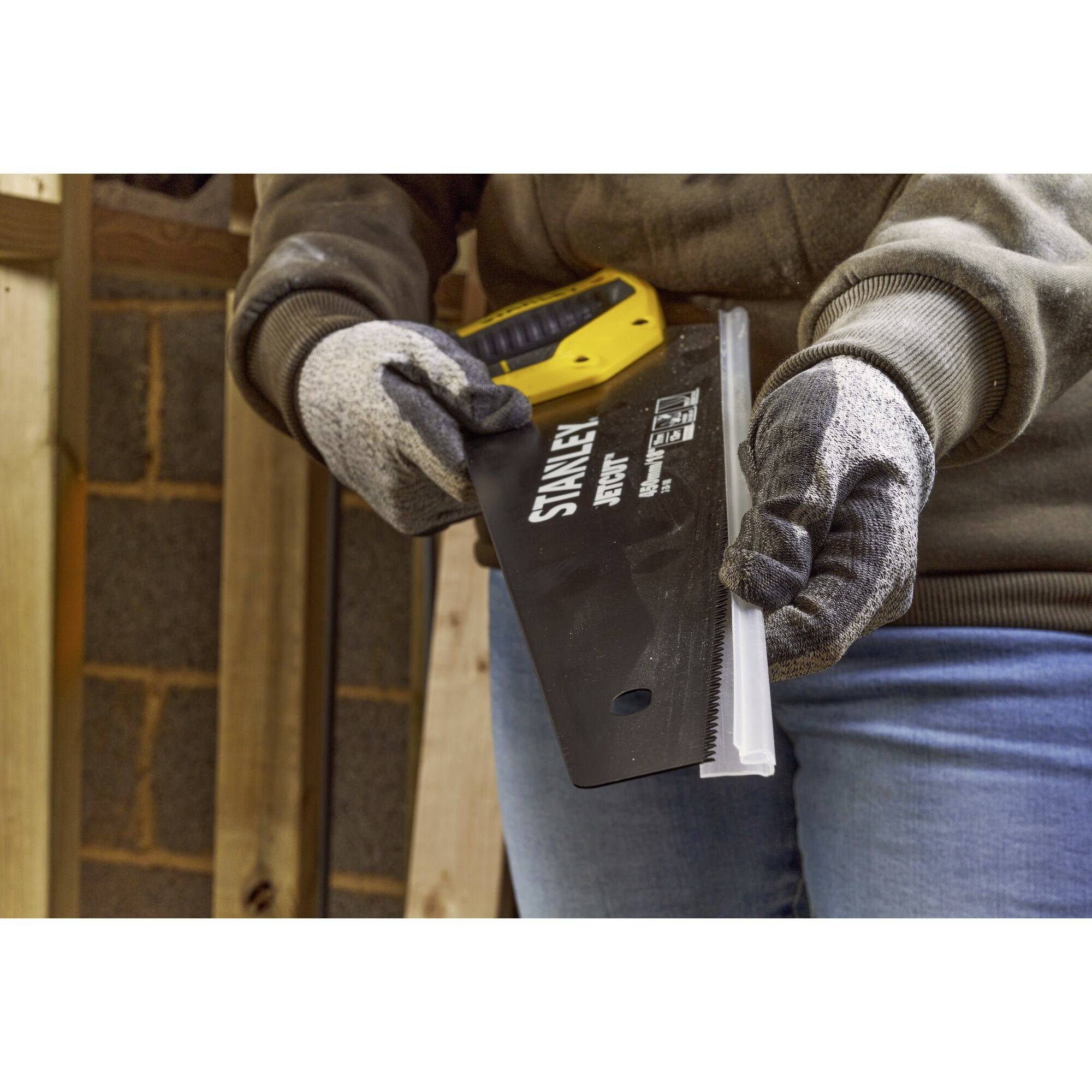 Osoba ubrana jest w rękawice i trzyma piłę ręczną, gotowa do cięcia drewna. Scena przedstawia rzemieślniczą pracę w warsztacie.