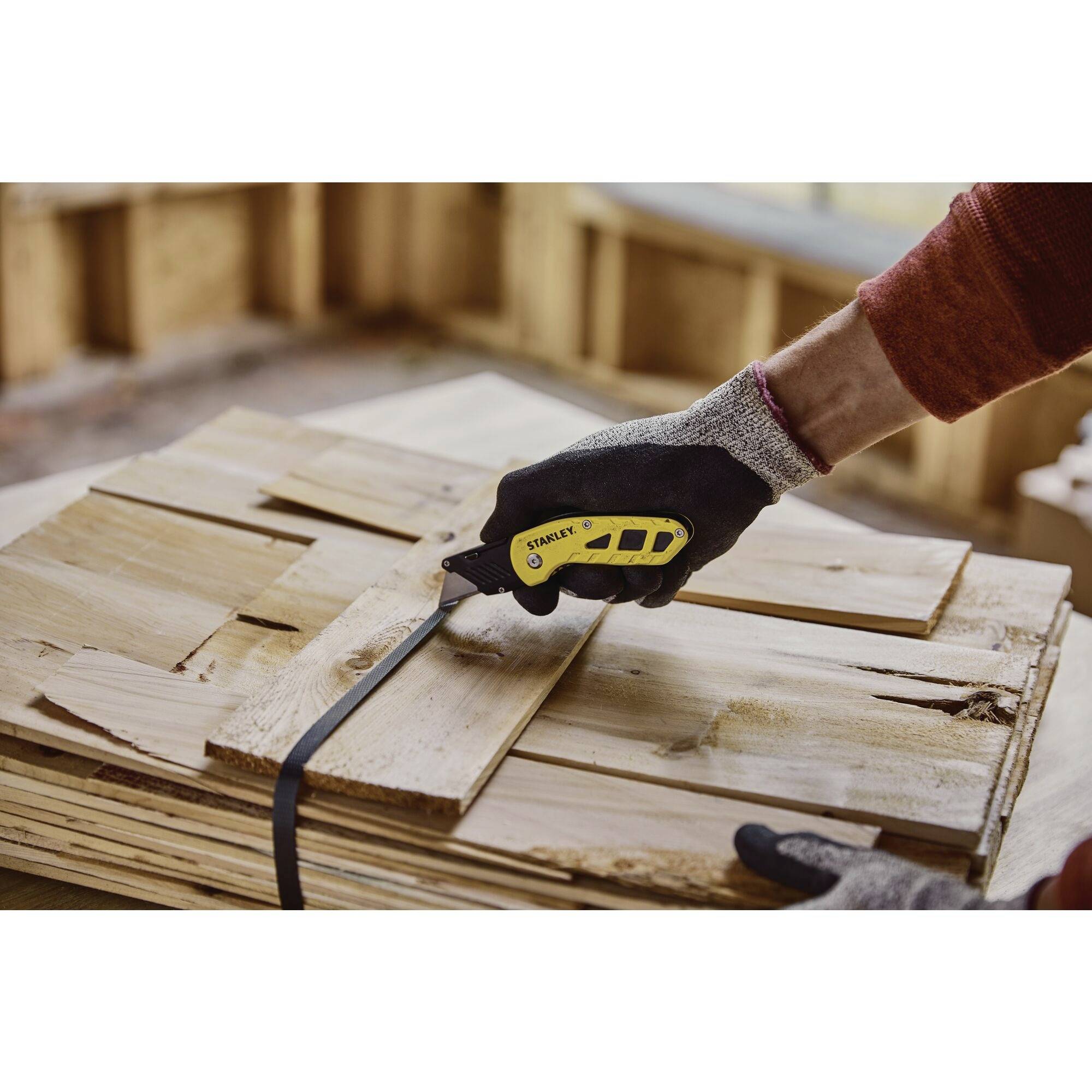 Osoba w rękawiczkach używa żółtego noża introligatorskiego do cięcia grubego kartonu na stosie drewna.