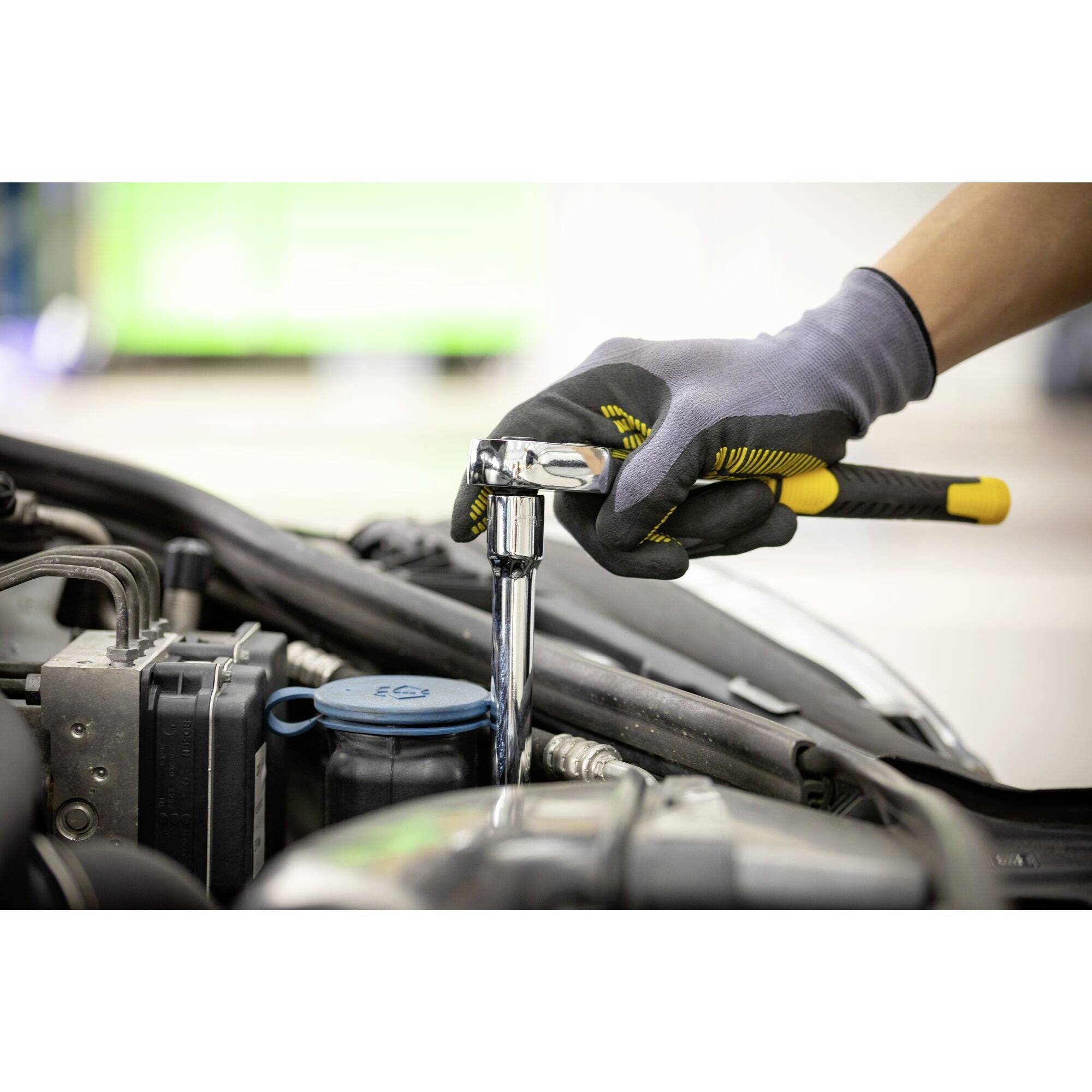 Ręka w rękawicy trzyma klucz mechaniczny i pracuje przy silniku samochodu. W tle widoczne środowisko warsztatu.