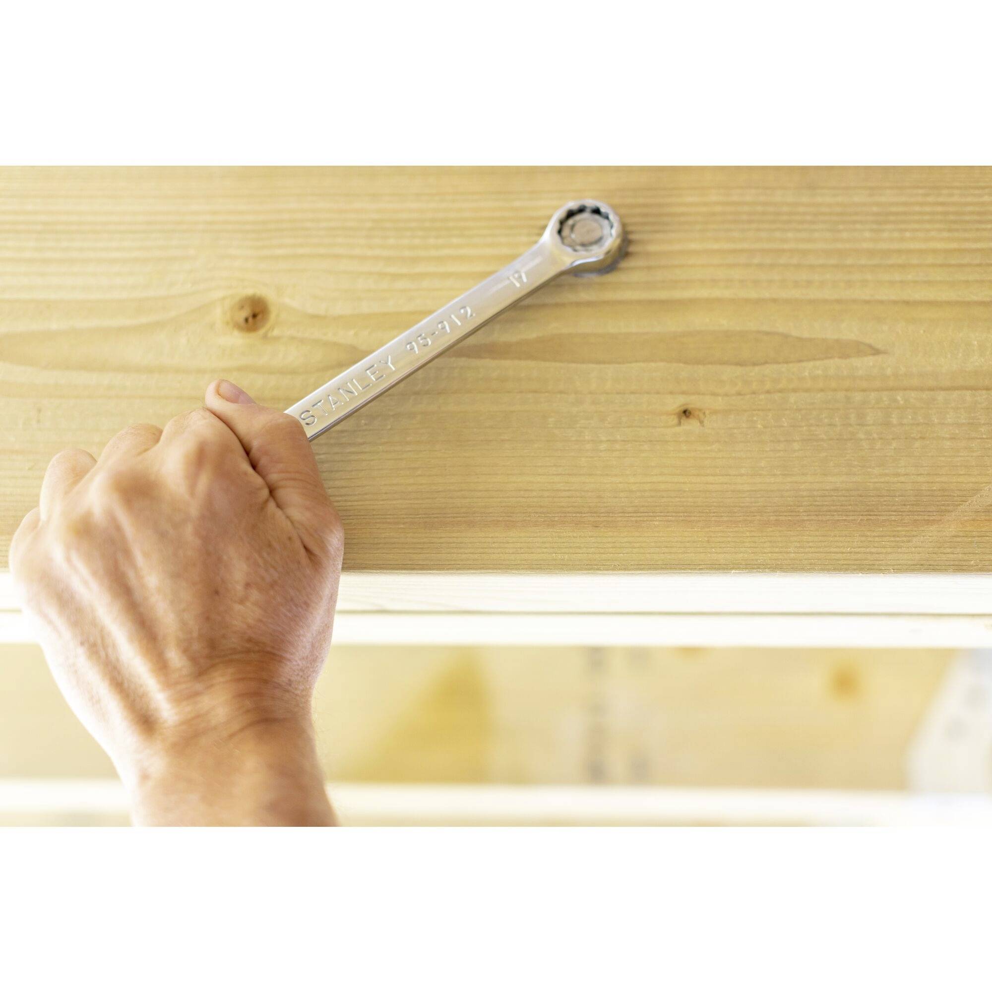 Dłoń trzyma klucz płaski, który wkręca śrubę w deskę drewnianą.