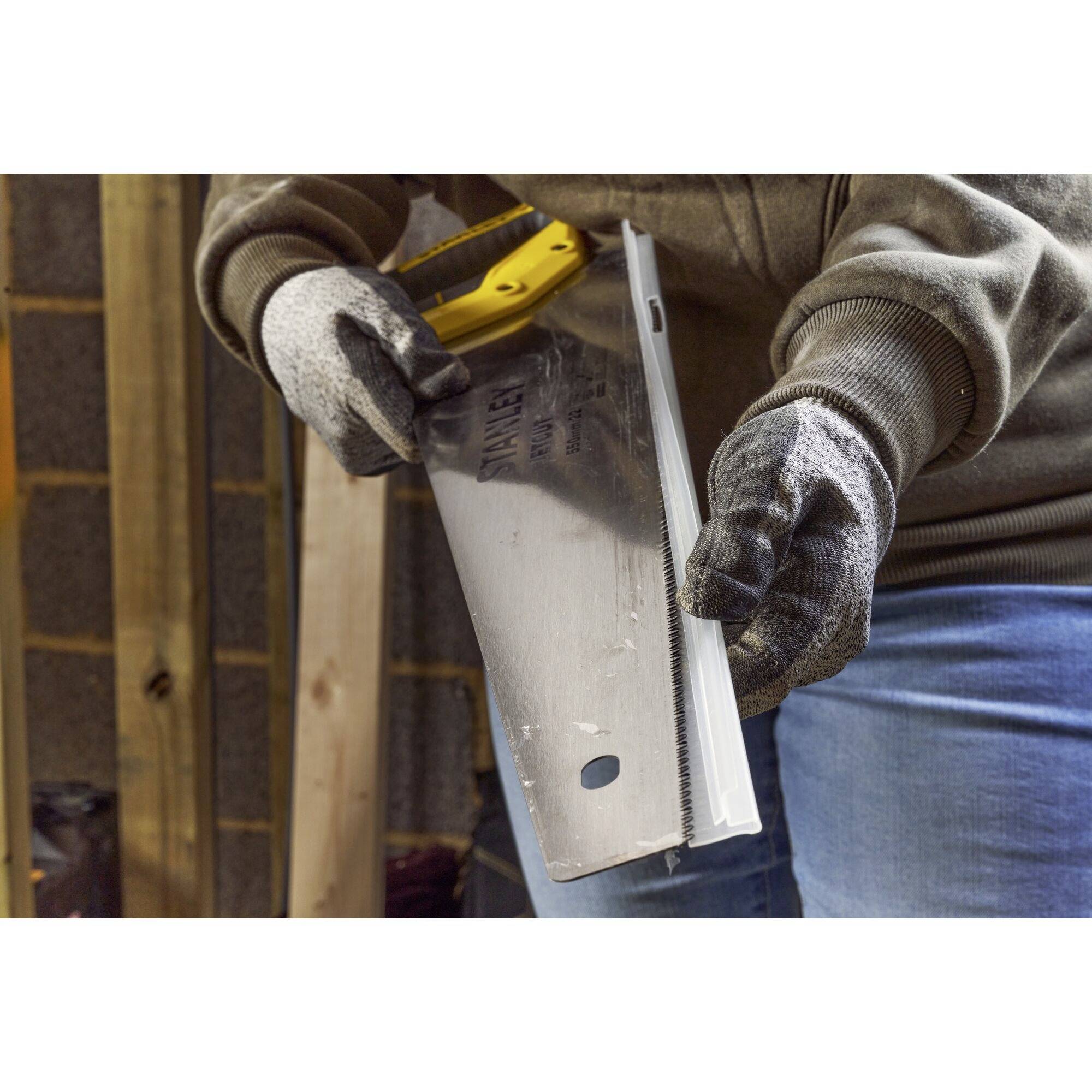 Osoba nosi rękawice i trzyma piłę ręczną, gotowa do cięcia. Otoczenie wskazuje na pracę rzemieślniczą lub projekt budowlany.