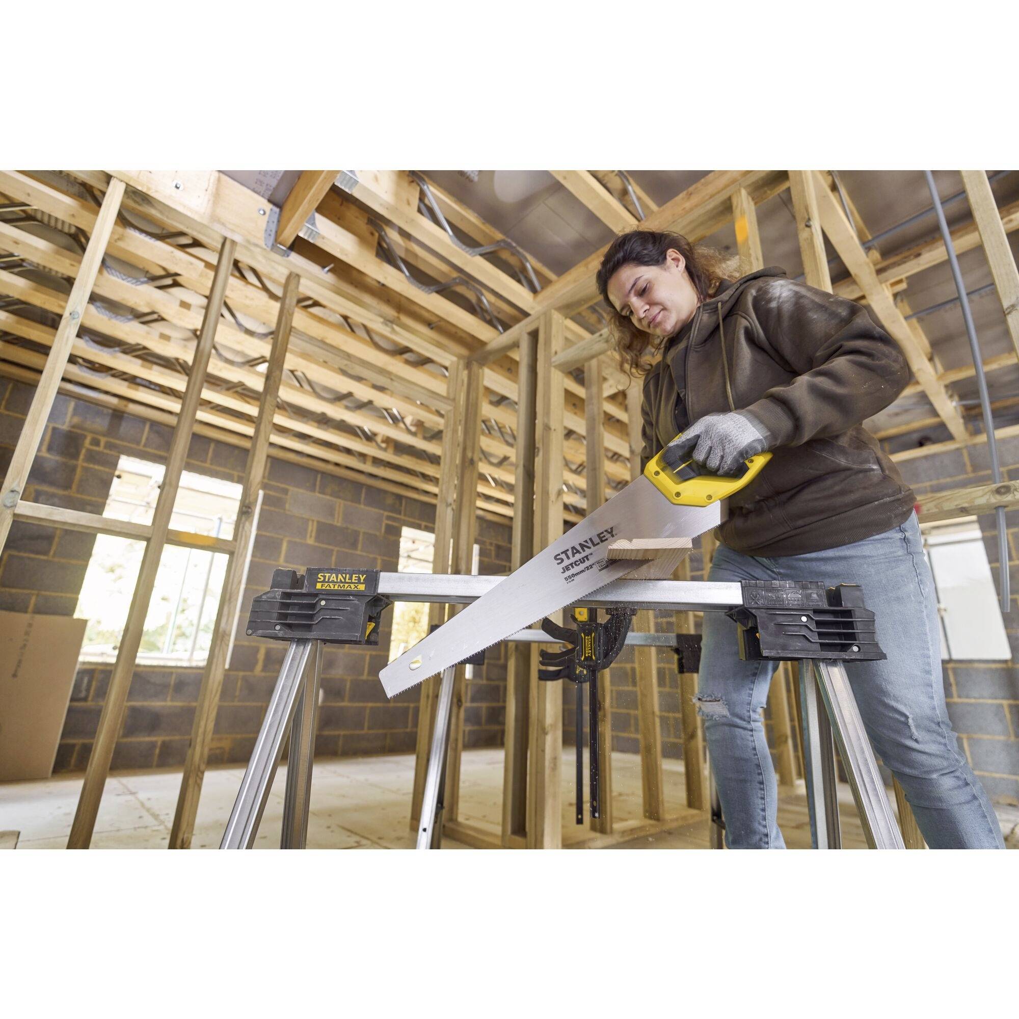 Osoba ubrana w strój roboczy używa piły ręcznej do cięcia drewna w pomieszczeniu będącym w trakcie budowy.