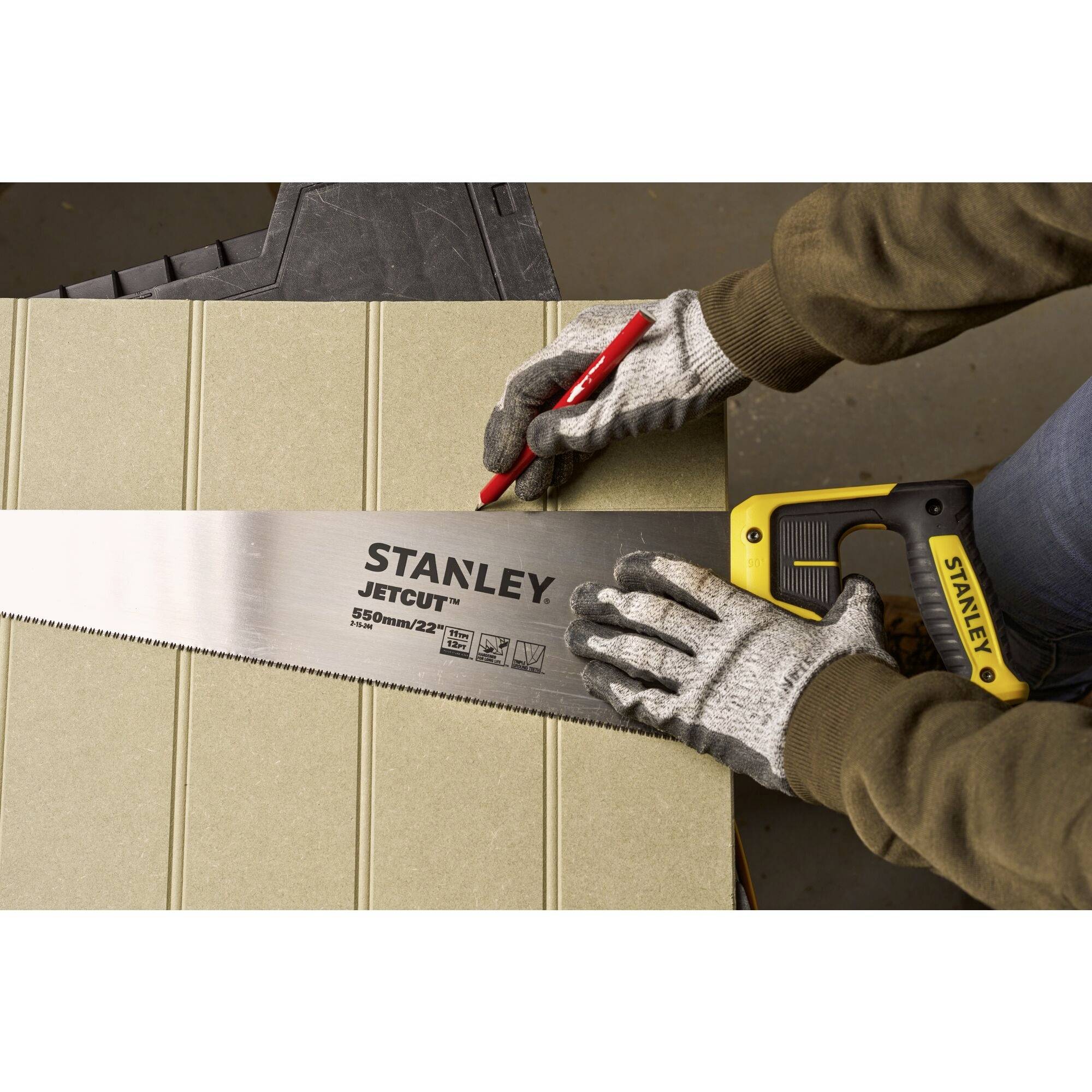 Osoba w rękawicach trzyma ręczną piłę Stanley i zaznacza deskę ołówkiem. Główny fokus: użycie narzędzia.