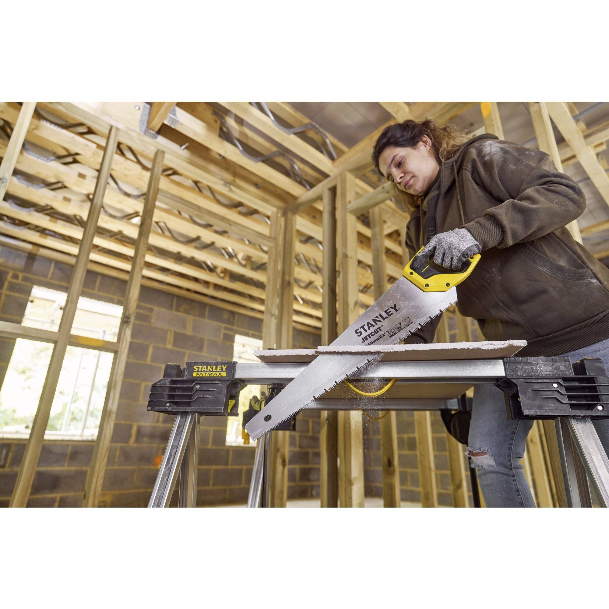Osoba przecina drewnianą deskę ręczną piłą na placu budowy. W tle widoczne są drewniane belki i murowane ściany.