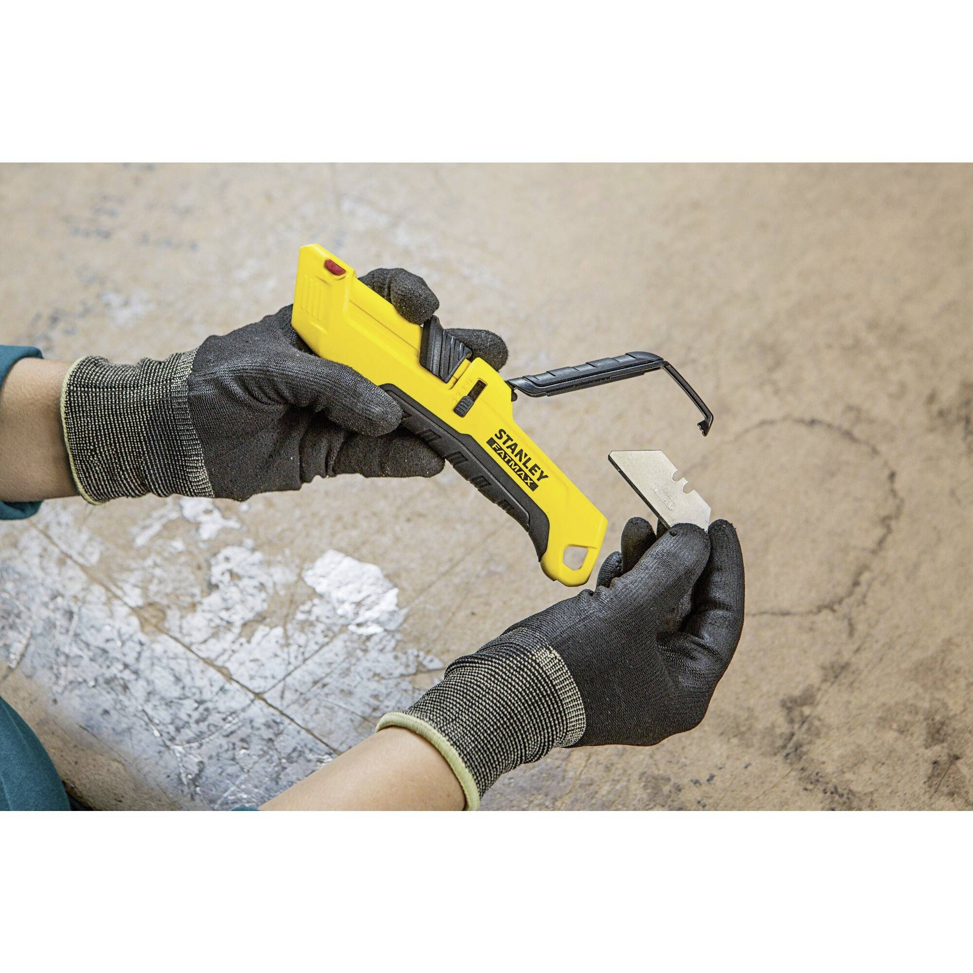 Obraz przedstawia dłonie w czarnych rękawiczkach trzymające żółty nóż roboczy z wymiennym ostrzem na szarym tle.