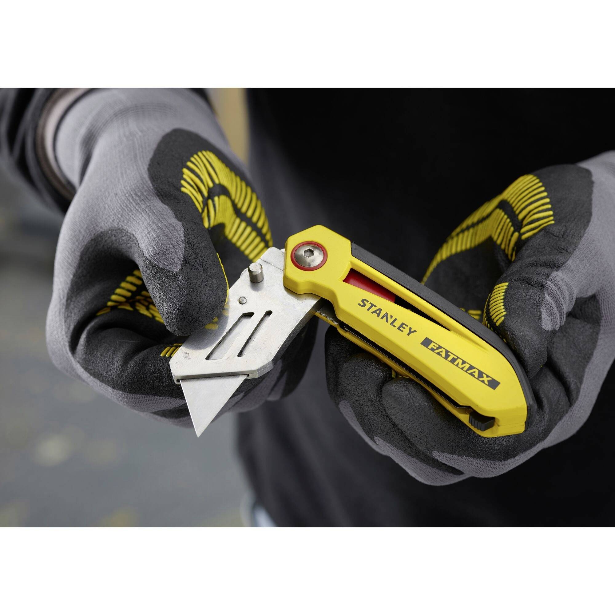 Osoba w rękawicach roboczych trzyma żółty nóż monterski Stanley Fatmax i wymienia ostrze.
