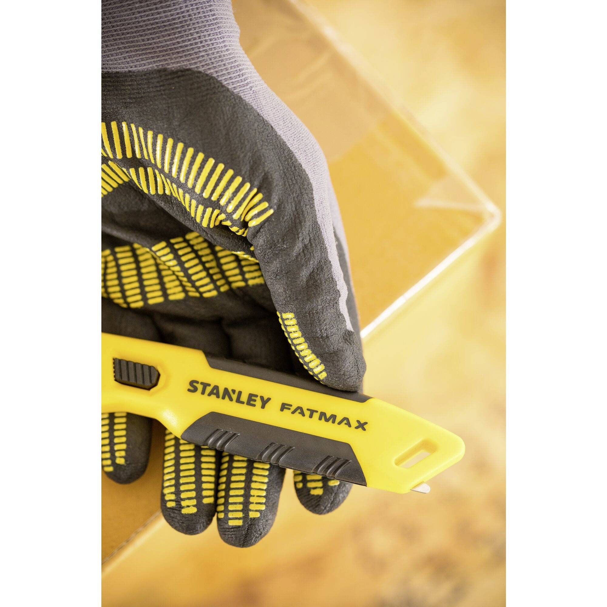 Dłoń w roboczej rękawicy trzyma żółty nóż monterski Stanley FatMax, który przecina karton.