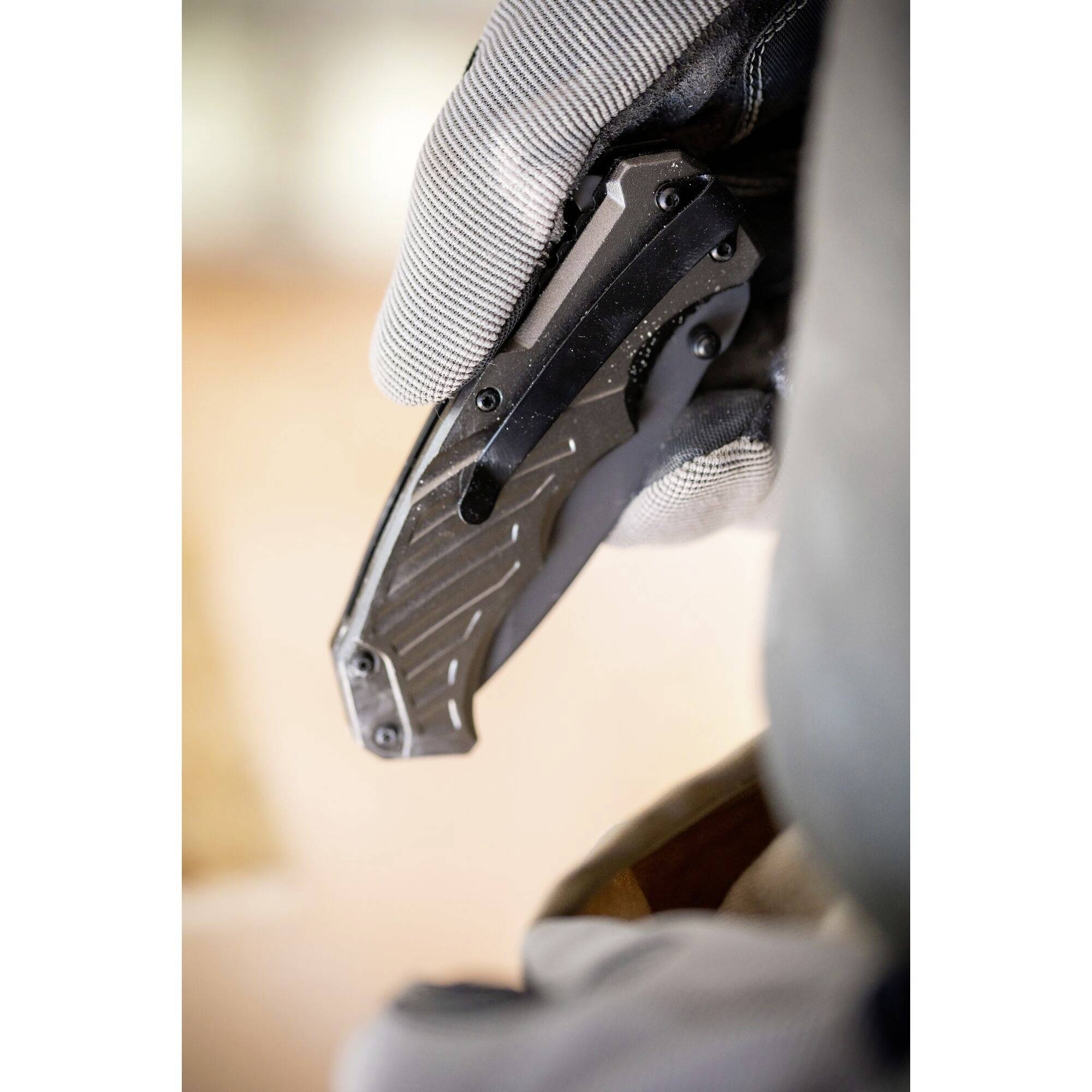 Dłoń w rękawiczce trzyma złożony czarny nóż składany. Tło jest rozmyte.