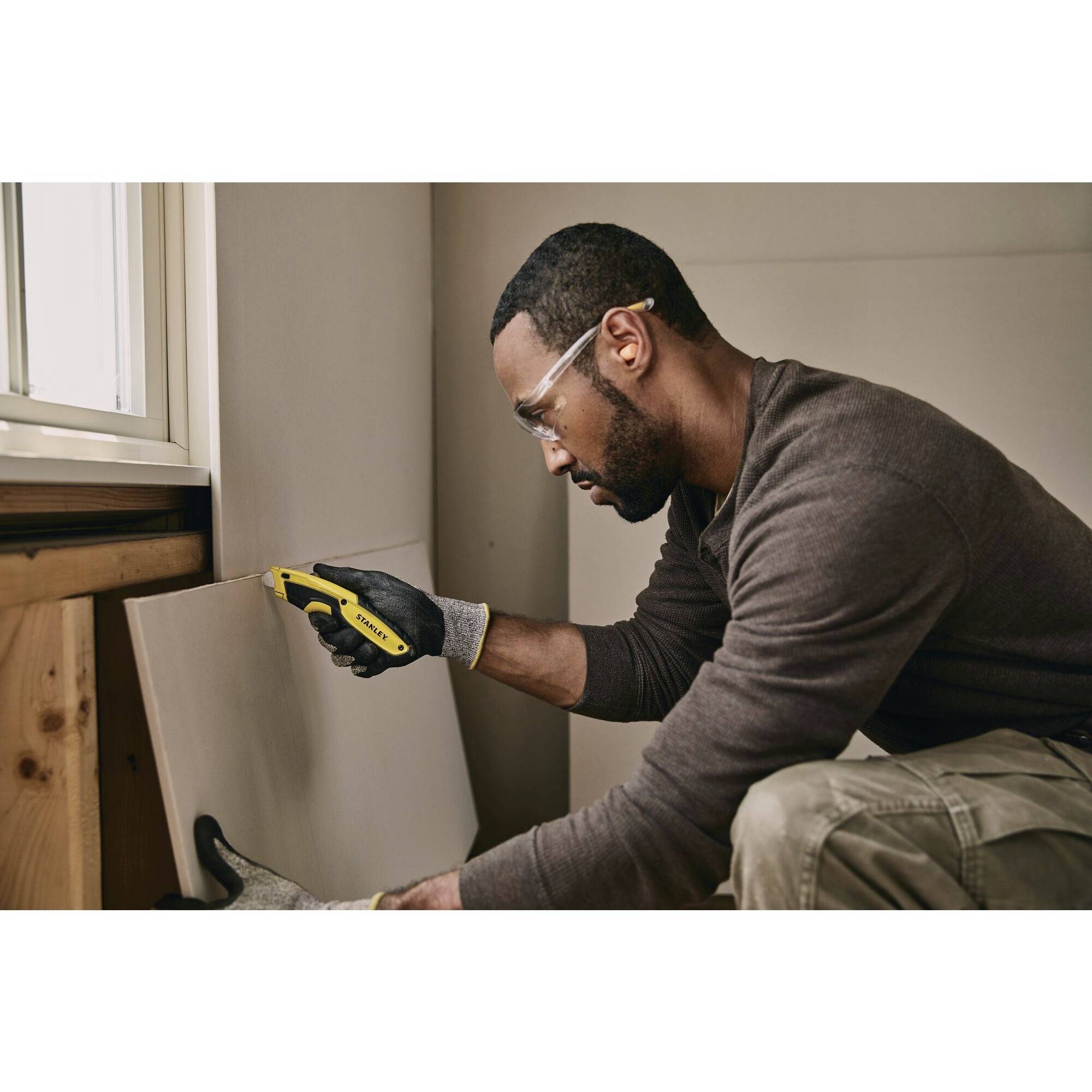 Osoba w okularach ochronnych i rękawicach przecina deskę drewnianą żółtym narzędziem we wnętrzu pomieszczenia.