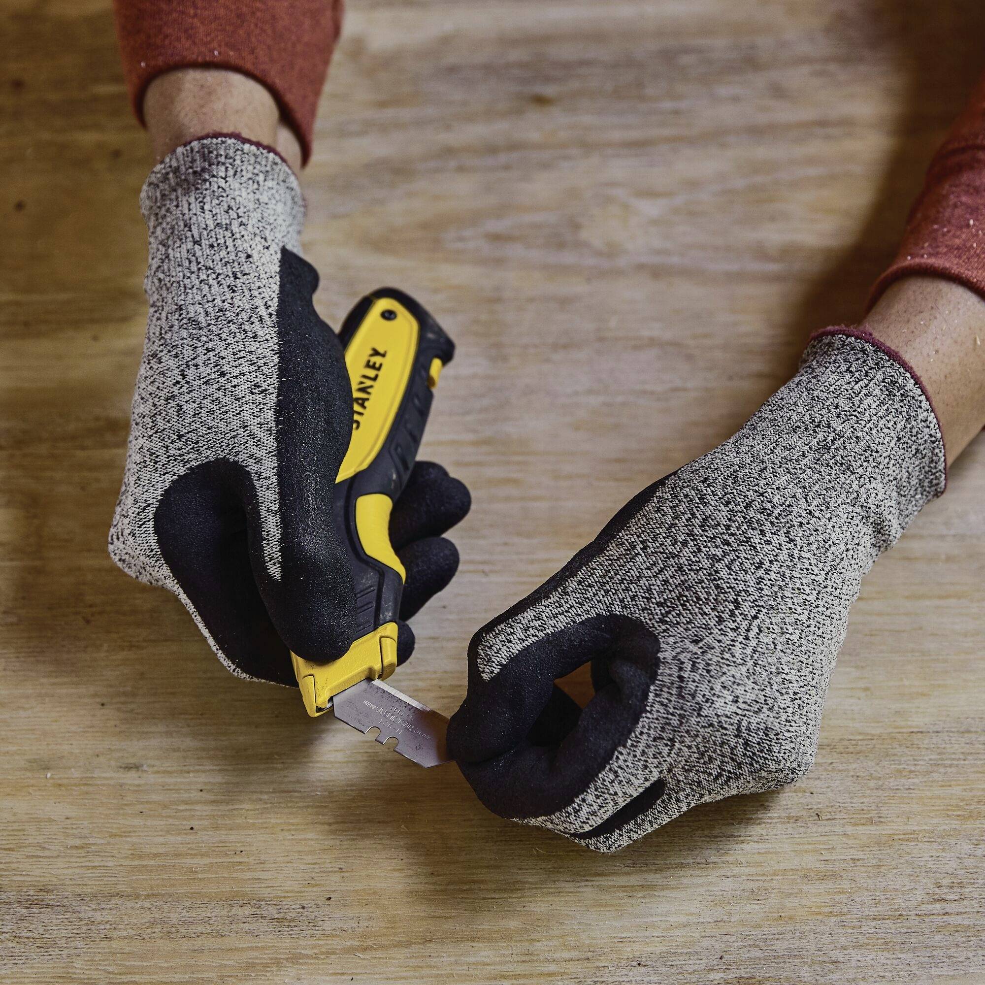 Para rąk w szarych rękawicach trzyma żółty nóż monterski podczas wymiany ostrza, na drewnianej powierzchni.