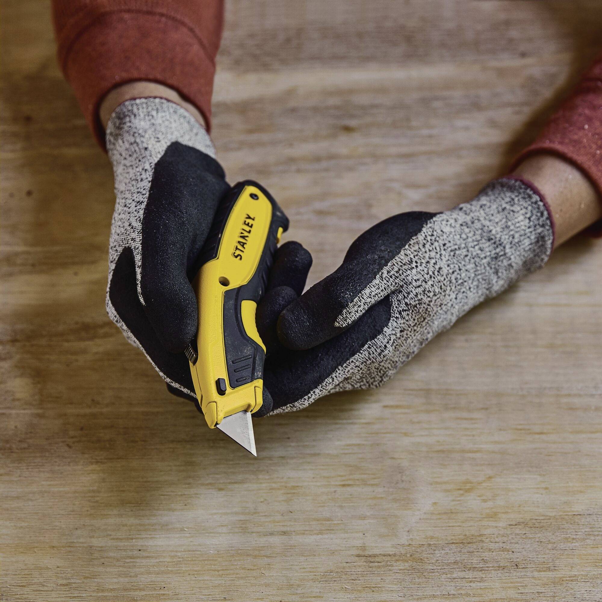 Osoba w rękawicach trzyma żółty nóż do dywanów nad drewnianym stołem, przygotowując go do użycia.